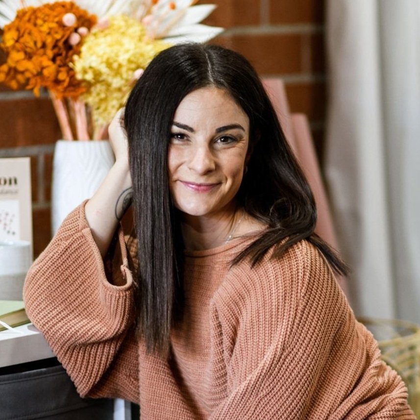 A woman with long dark hair, smiling and winking, wearing a brown sweater, sitting indoors with a vase of orange and yellow flowers in the background.