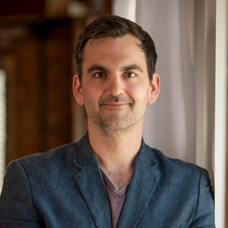 A man with short dark hair, light beard, wearing a dark blazer and a maroon V-neck shirt, standing indoors near a window with curtains, smiling softly.