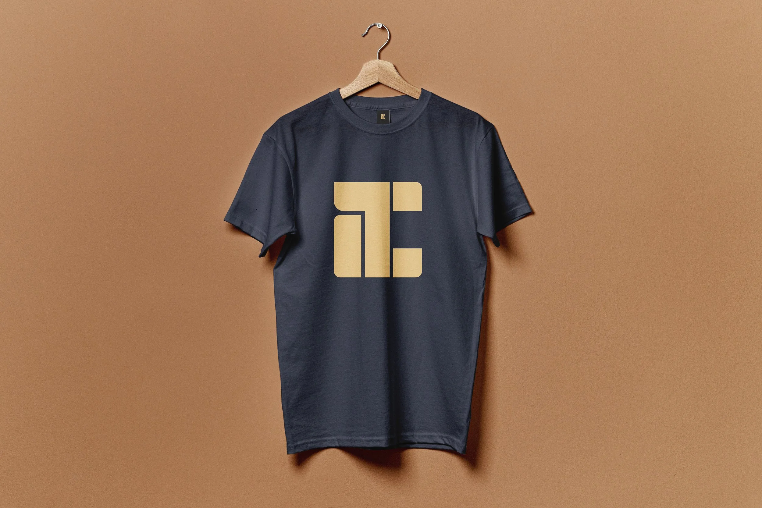 A dark gray T-shirt hanging on a wooden hanger on a tan wall, with a large beige logo in the center of the chest area that resembles the letter 'E' composed of geometric shapes.