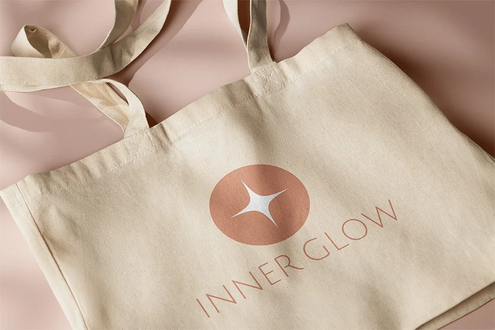 Cream-colored tote bag with the Inner Glow logo, a circle with a star-shaped design, printed on the front, lying on a light pink surface.