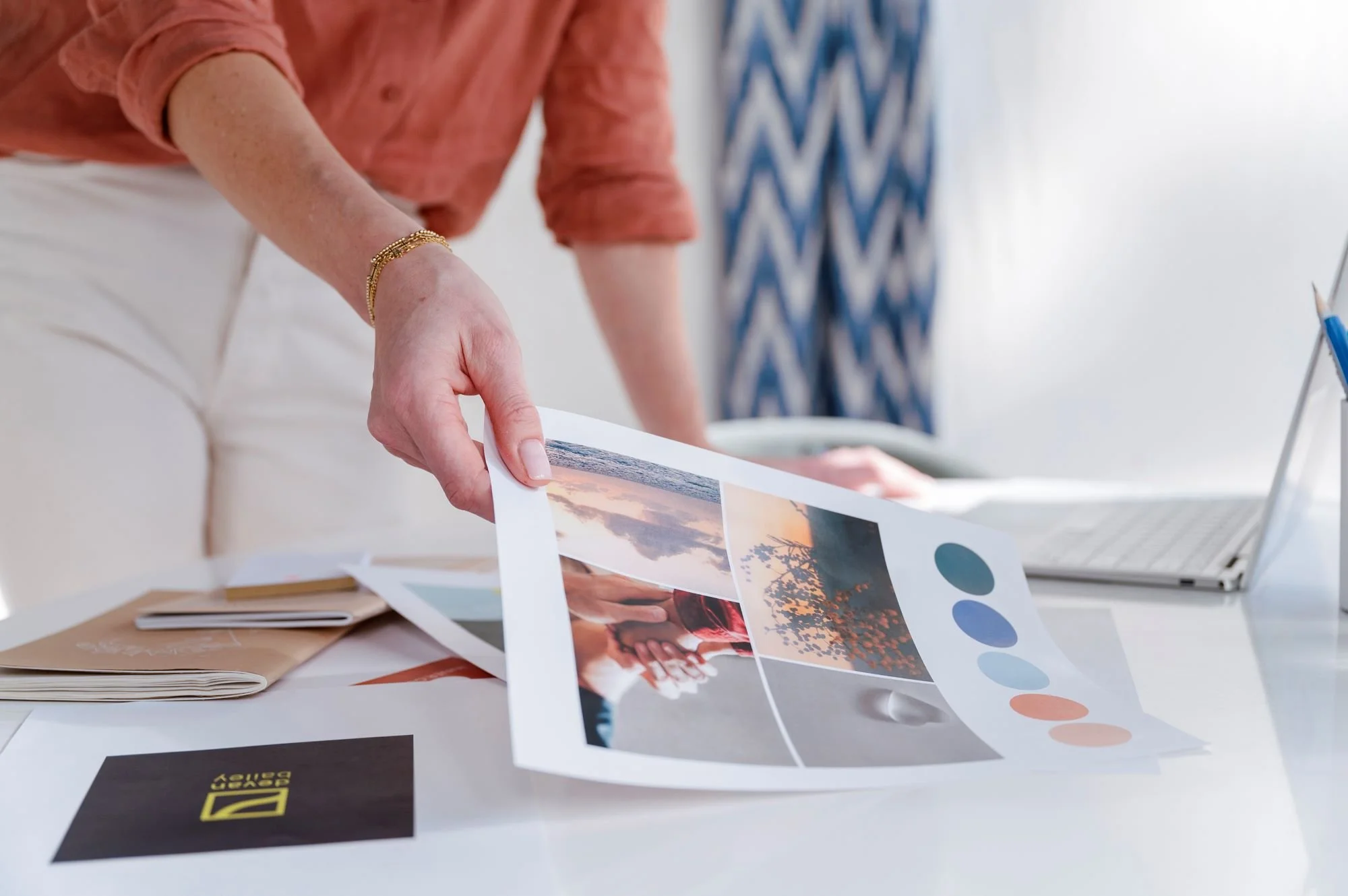 A person in a peach shirt and cream pants is at a desk, holding a printed photograph or artwork, with a laptop, color swatches, and papers also on the desk. The background shows a blurred patterned curtain.