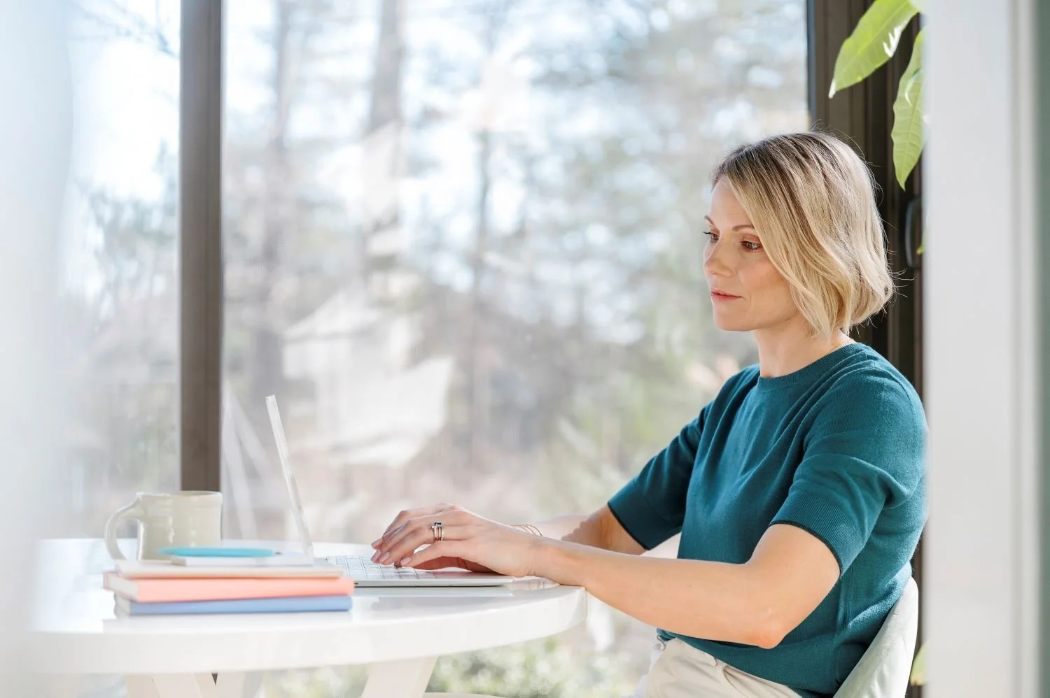 A woman with blonde hair working on a laptop at a white table by a large window, with a mug and closed notebooks on the table.