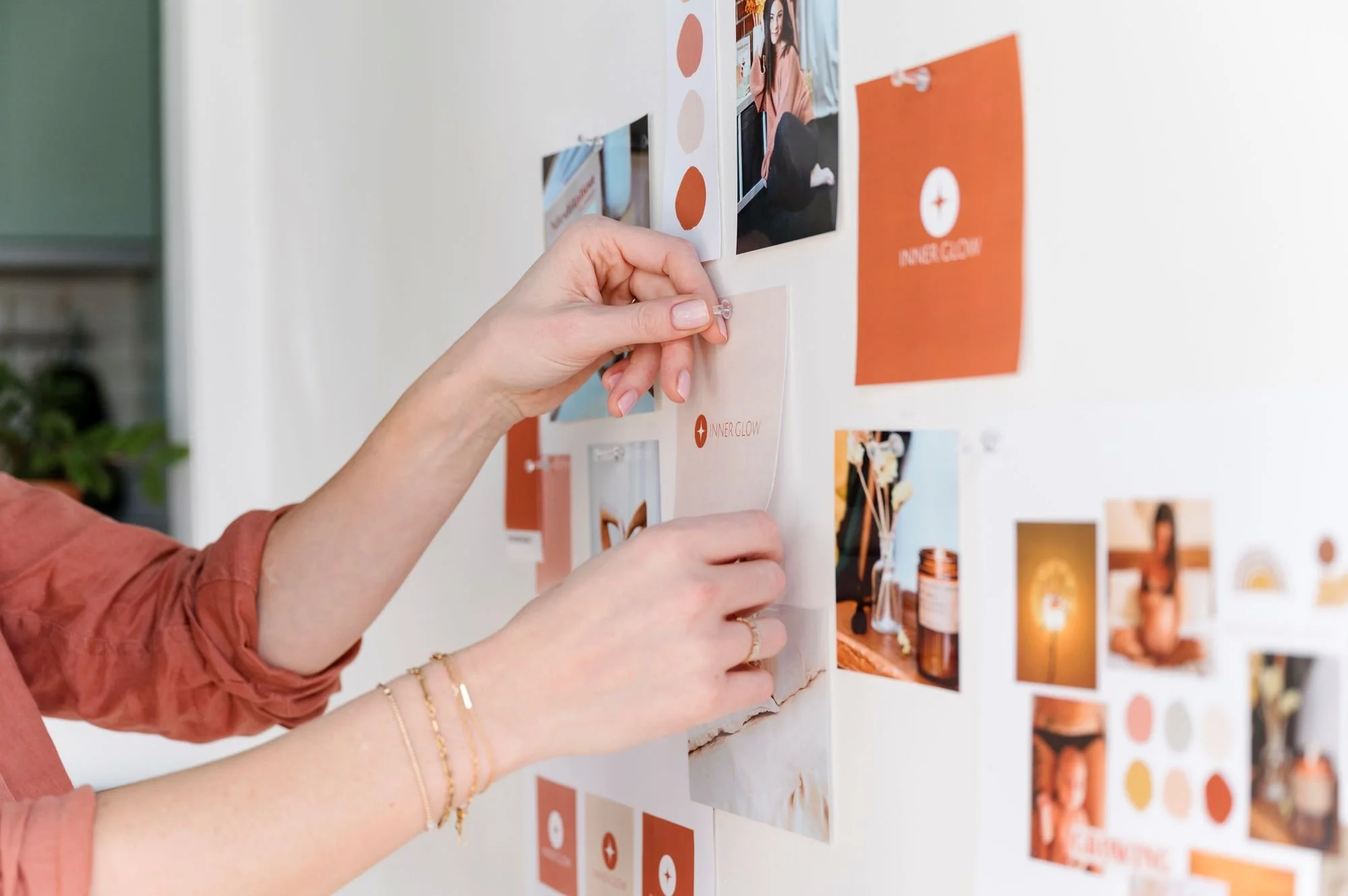 A person pinning a photo onto a white wall with other photos and color swatches, some featuring branding with the words 'Inner Glow'.
