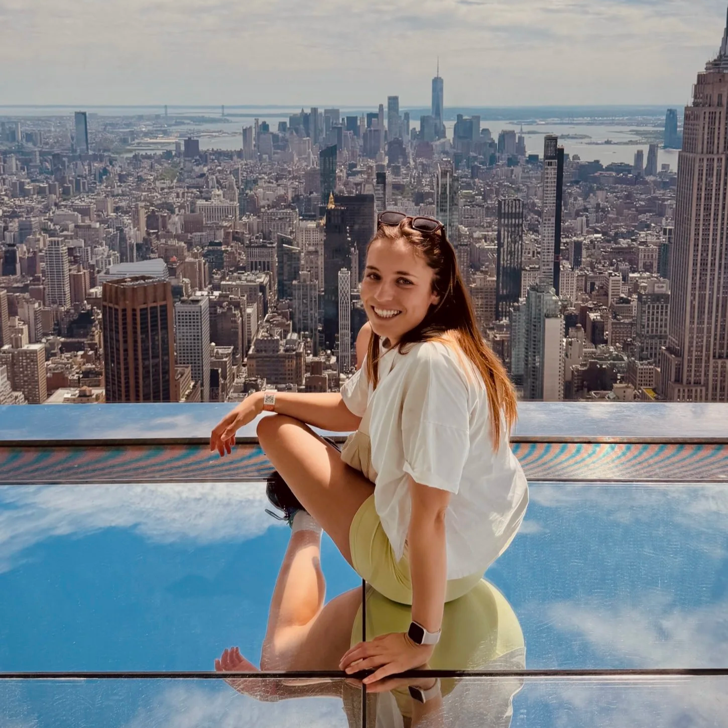 Eine lachende Frau sitzt auf einem reflektierenden Ball vor der Skyline von New York City, mit vielen Wolkenkratzern und Blick auf den Fluss im Hintergrund.