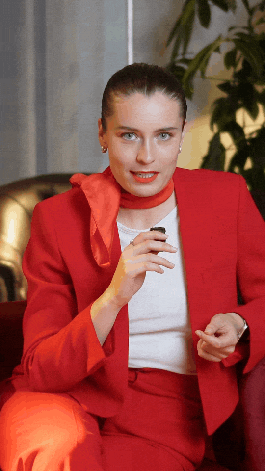 Eine Frau in rotem Anzug und halstuch sitzt auf einem Sofa, hält eine Zigarette in der Hand, im Hintergrund sind Pflanzen und eine goldene Sofa-Armlehne sichtbar.