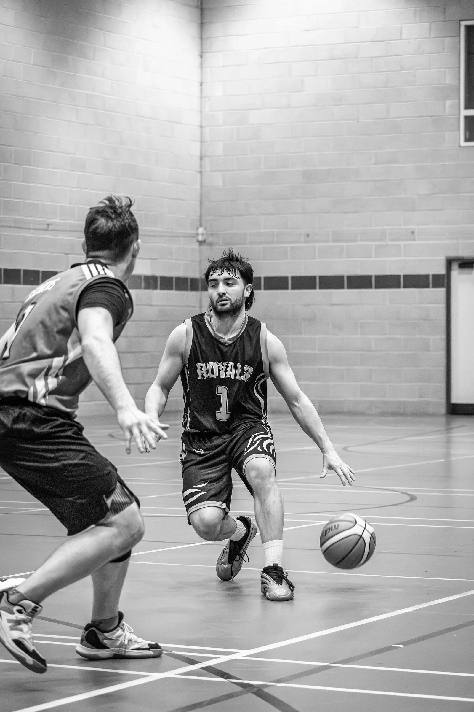 Two basketball players in action on an indoor court, one dribbling the ball and the other defending, both wearing jerseys and athletic shoes.