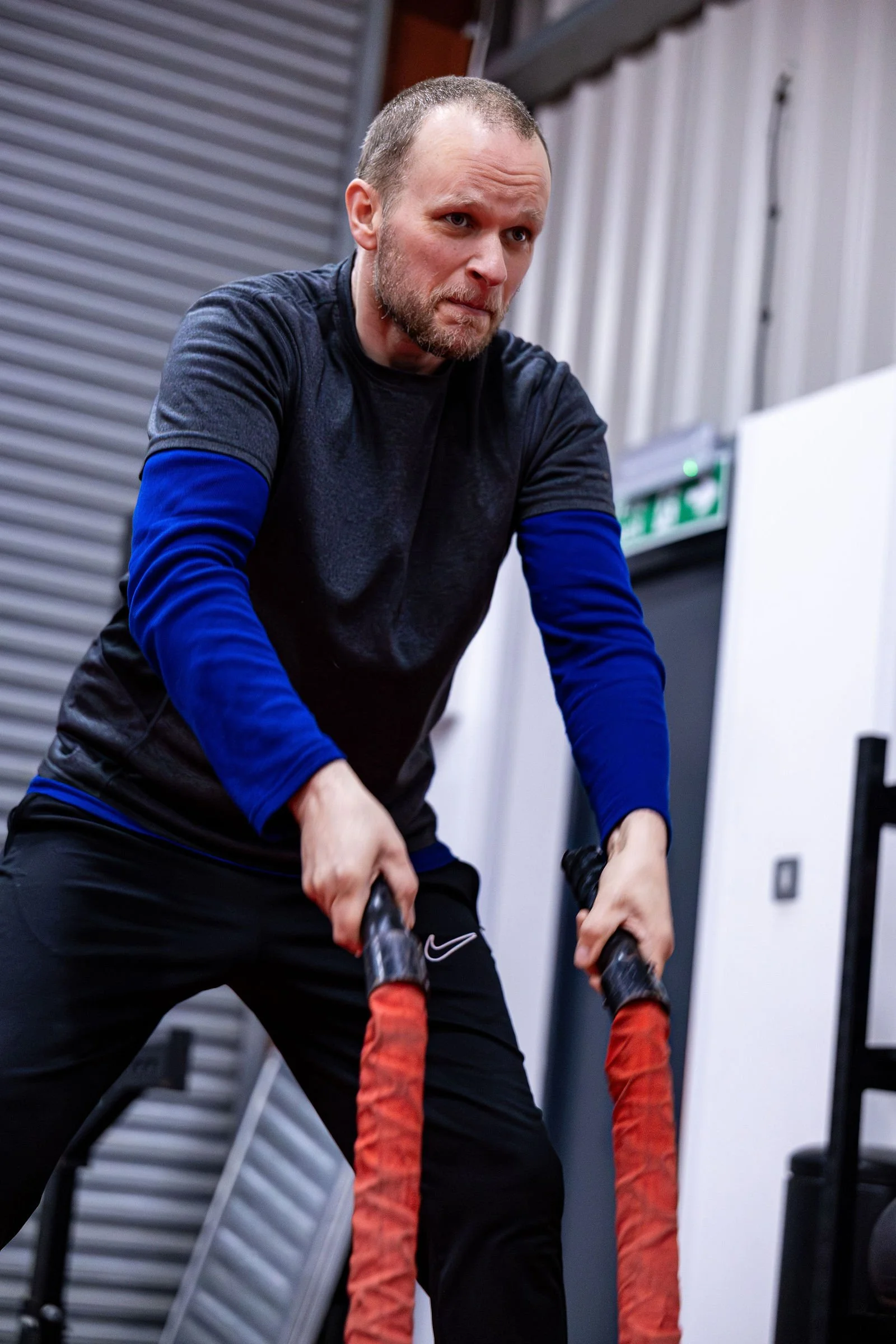 A man with a beard and short hair, wearing a black shirt with blue sleeves, working out with battle ropes in a gym.