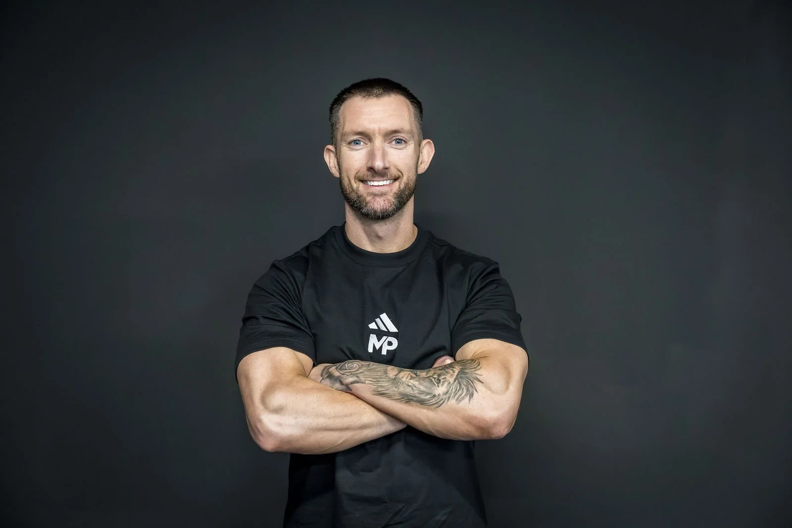 A smiling man with a beard and short hair, wearing a black sports shirt with a logo, standing with arms crossed against a dark background.
