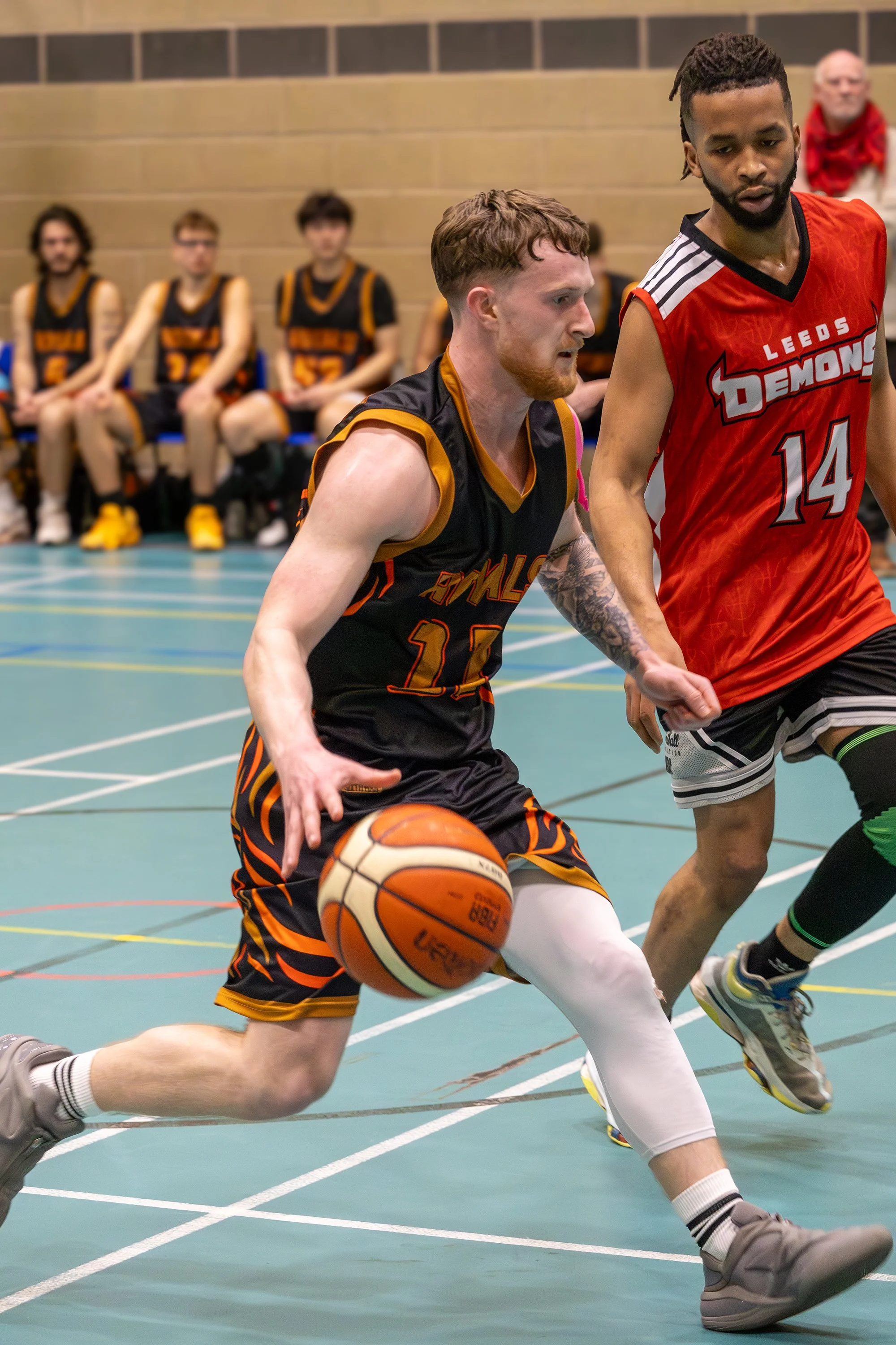 Two basketball players chase after a loose ball on the court during a game, while other players and a person in a red hoodie watch from the benches in the background.