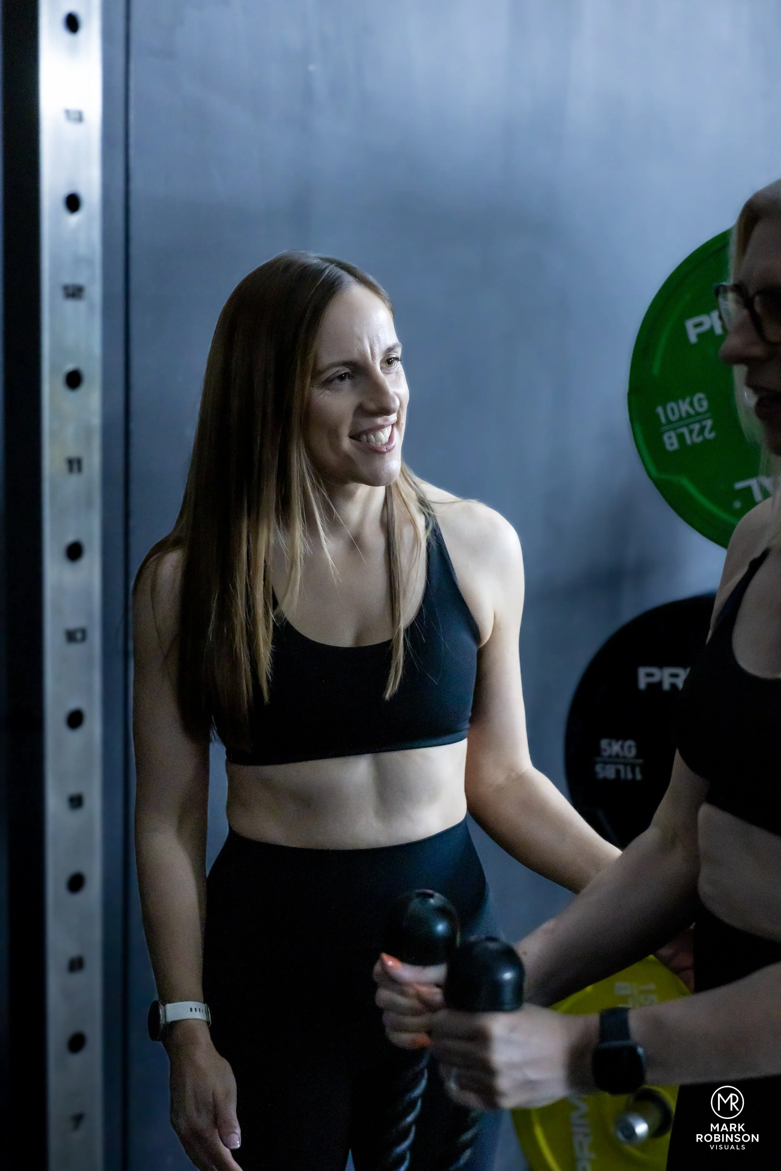 A fit woman with long brown hair wearing a black sports bra and leggings, smiling while chatting with another woman at the gym. The other woman is holding a water bottle and a protein shaker, and is wearing glasses with part of her face visible.