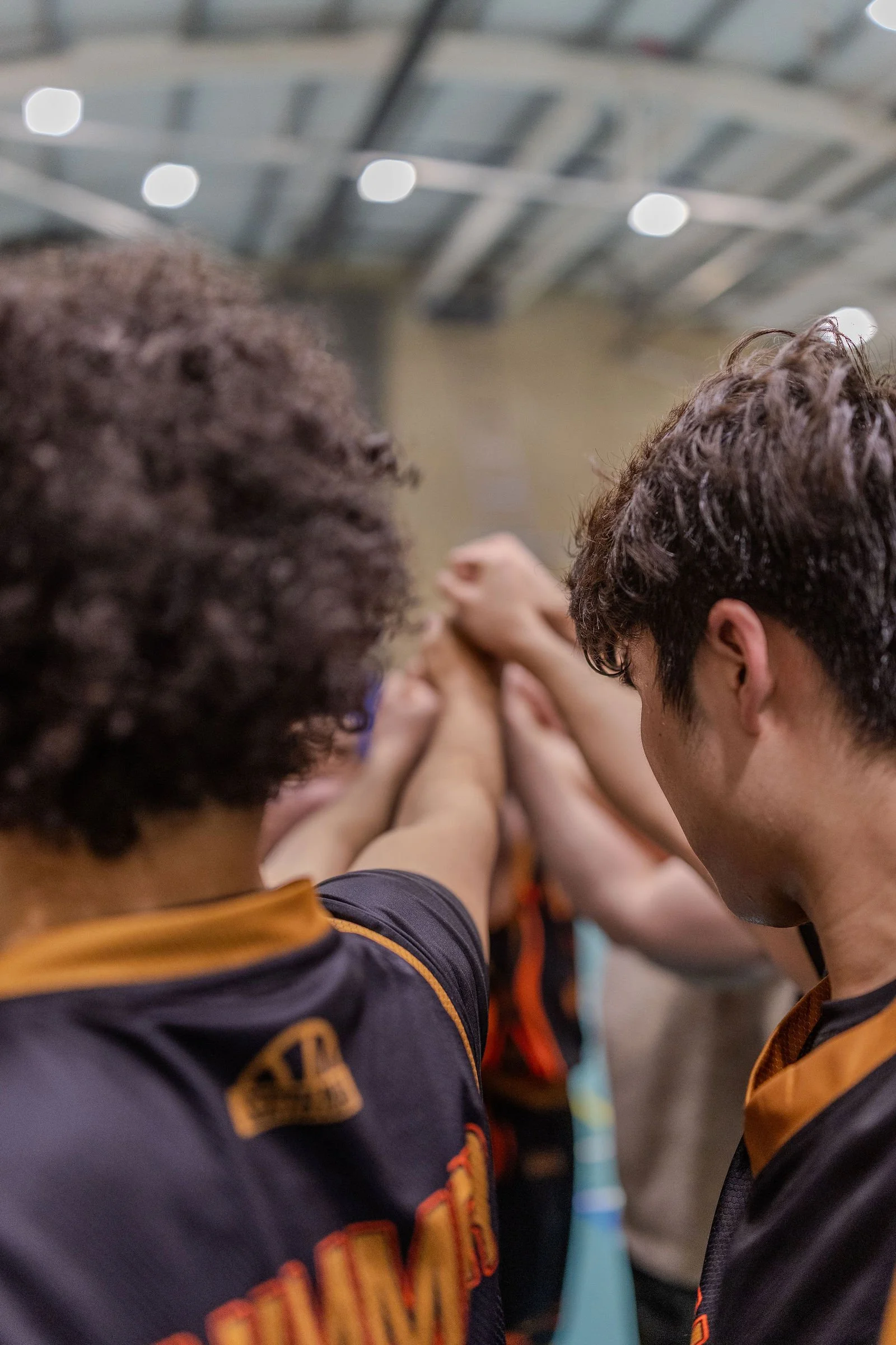 A group of people participating in a team huddle, with hands stacked together in the center, inside a gymnasium or similar indoor sports facility.