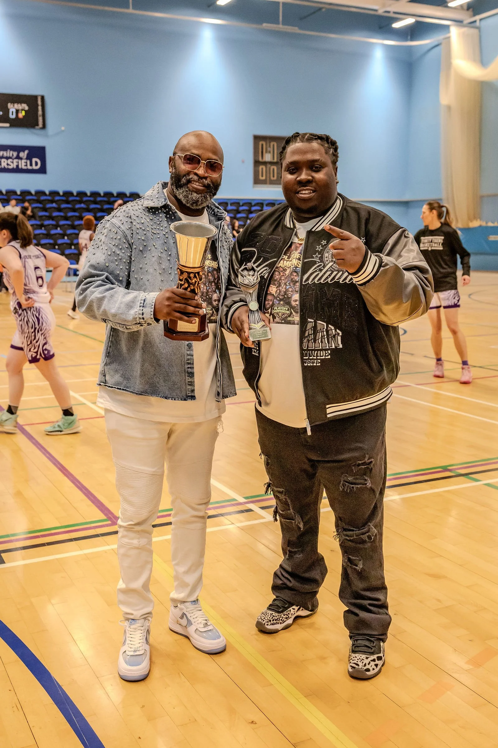 Two men standing on a basketball court holding trophies, with basketball players in the background.