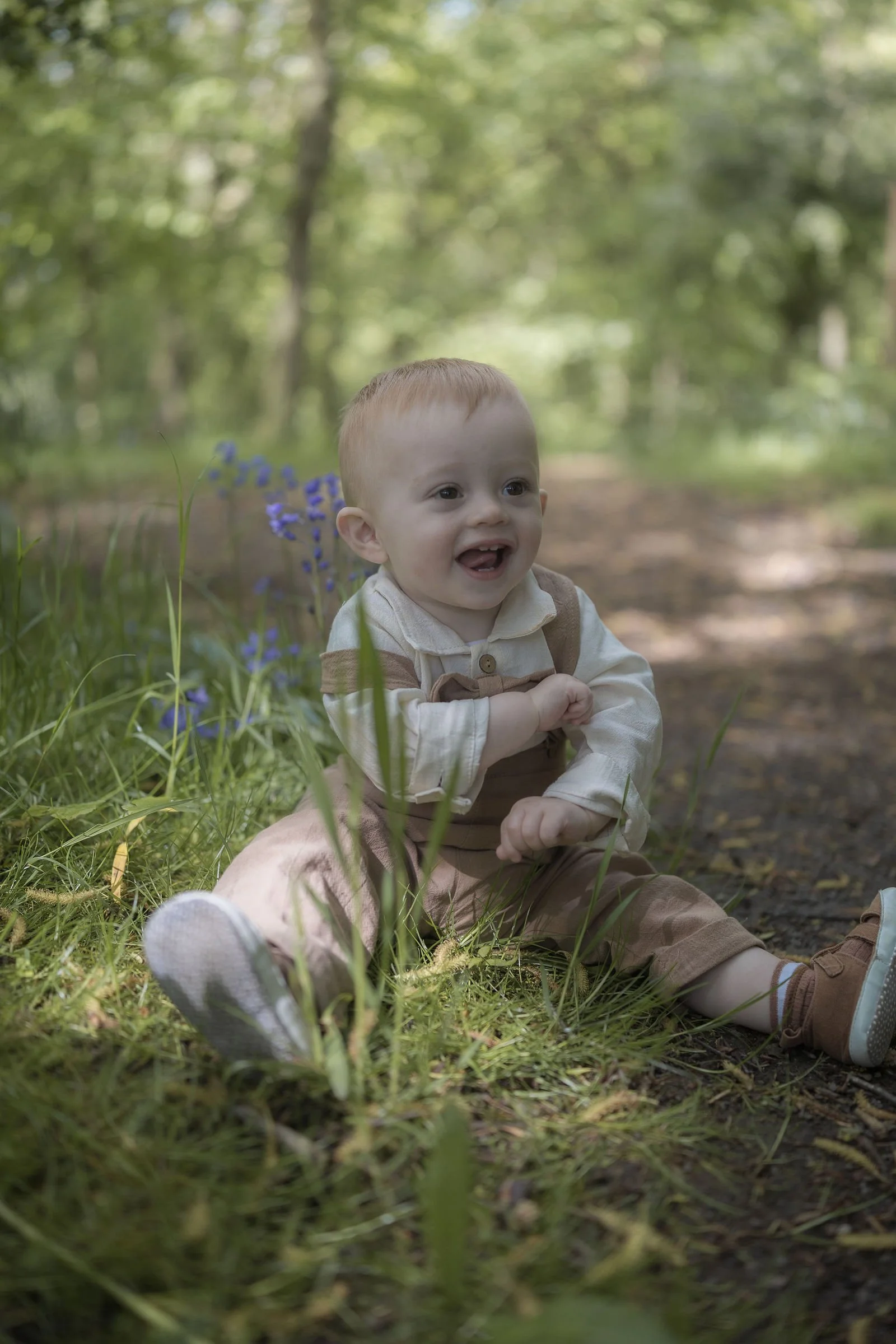 A young boy with light skin and red hair sitting outdoors on a forest trail, smiling with his tongue slightly out, surrounded by green trees and purple flowers.