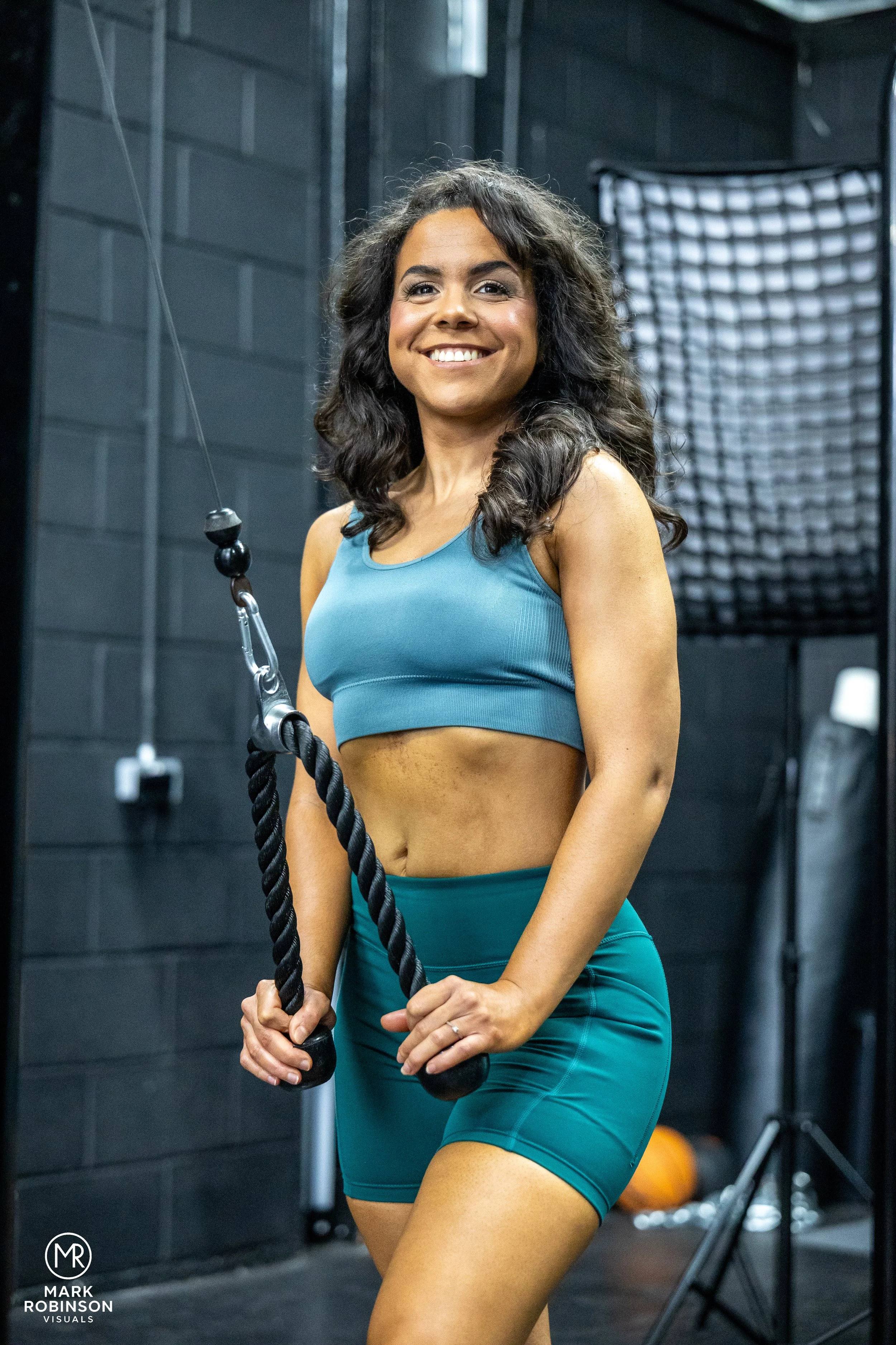 A woman in workout clothes holding a thick black rope in a gym, smiling at the camera.