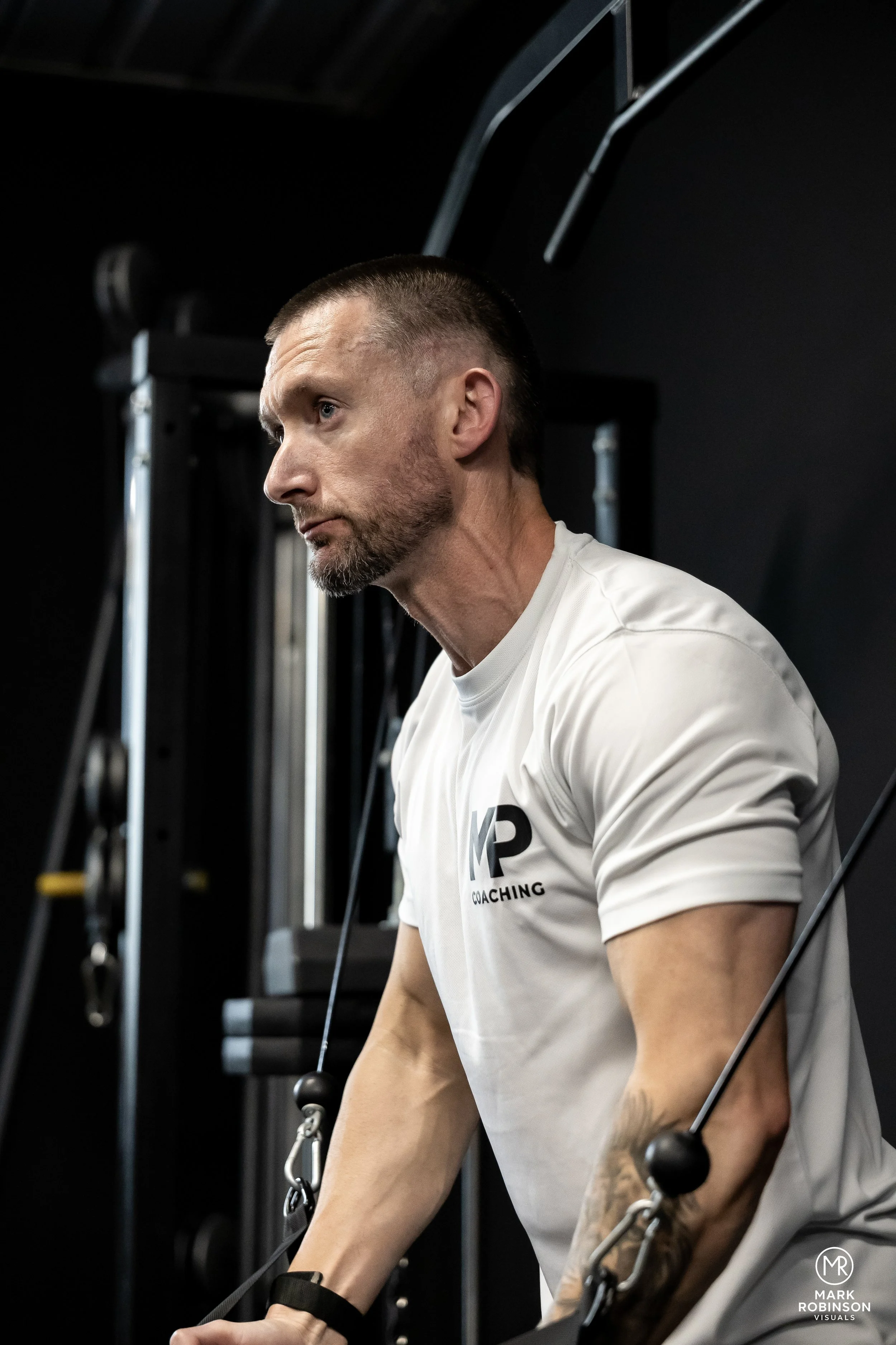 A man with short hair and a beard, wearing a white t-shirt with the word 'COACHING' on it, is working out at a gym using a cable machine. He appears focused and is in a side profile view.