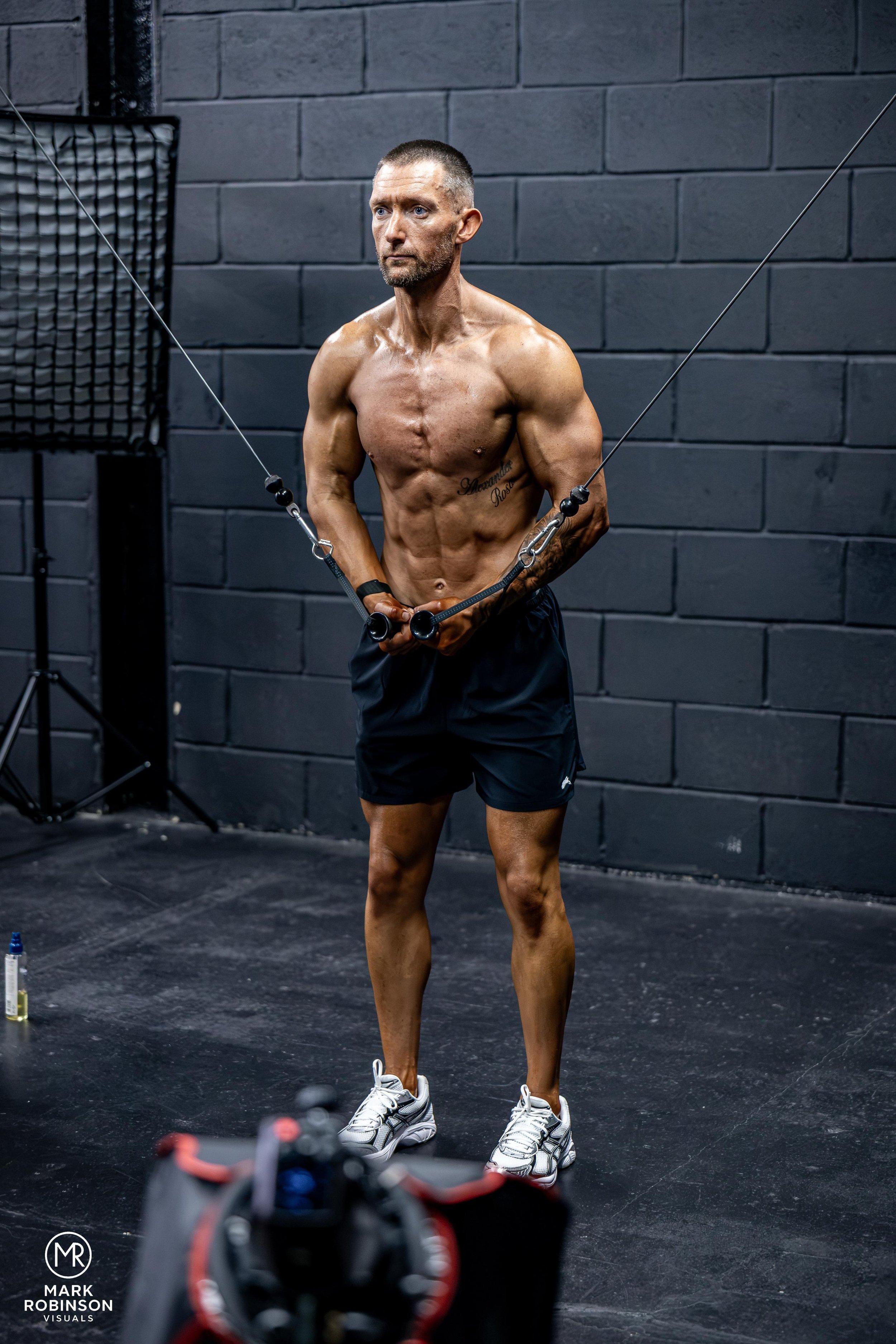 Michael Purdie with muscular build, tattoos, and a buzz cut stands in a gym against a dark brick wall, holding resistance bands. He is wearing black shorts and white sneakers. Equipment and a water bottle are on the floor nearby.