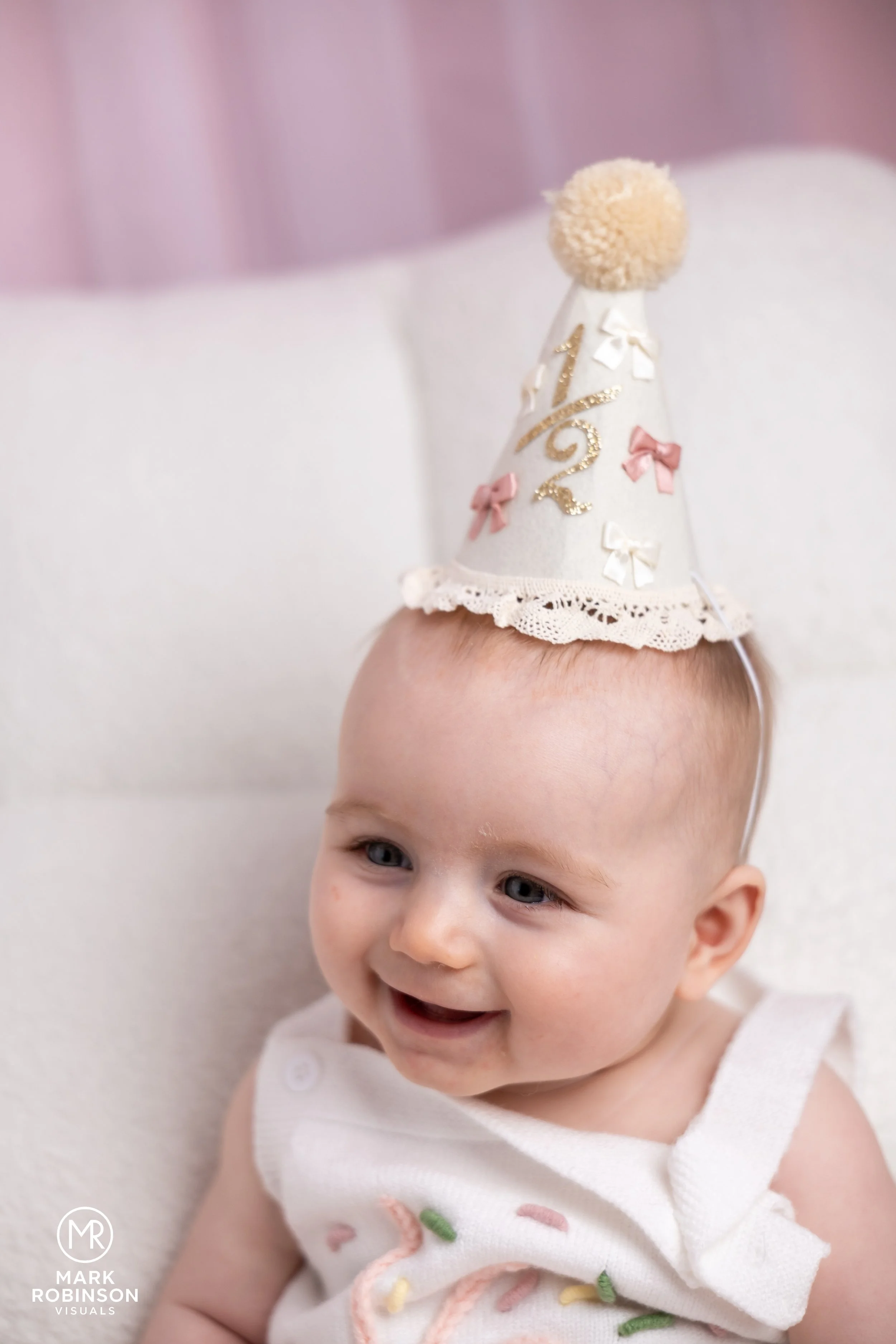 A smiling baby wearing a white sleeveless dress with colorful embroidery, and a birthday hat with pink and white bows and a pom-pom on top.