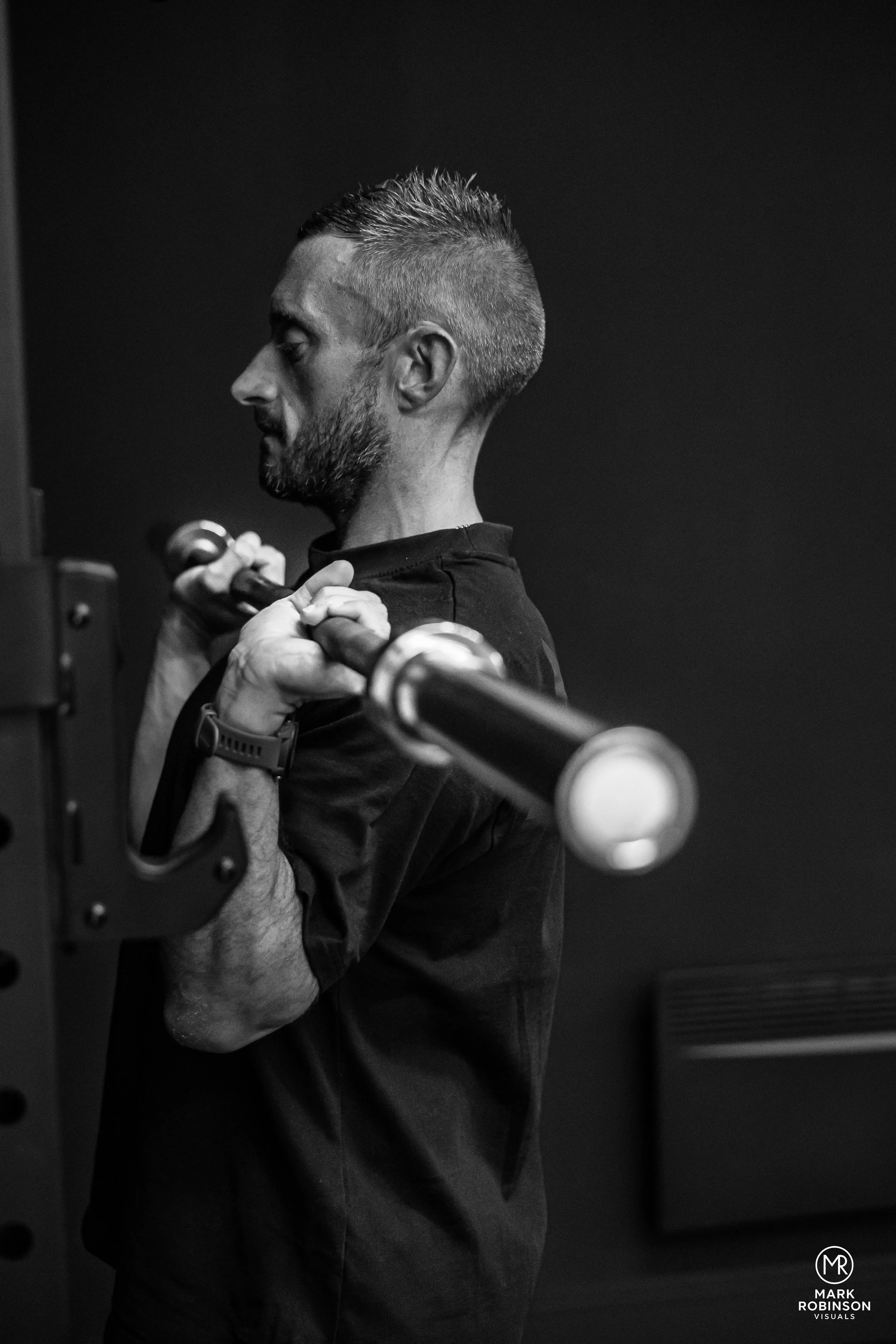 A man with a beard lifting a dumbbell in a gym. The image is in black and white with a plain dark background.