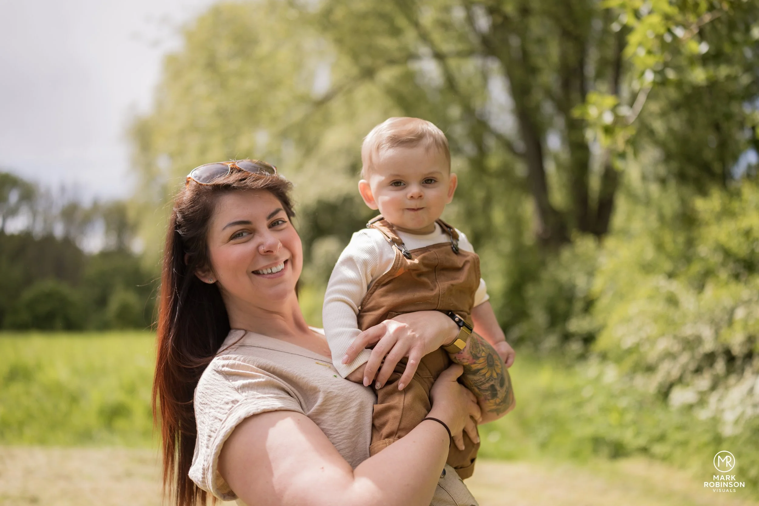 A woman holding a young boy outdoors in a park with trees in the background.