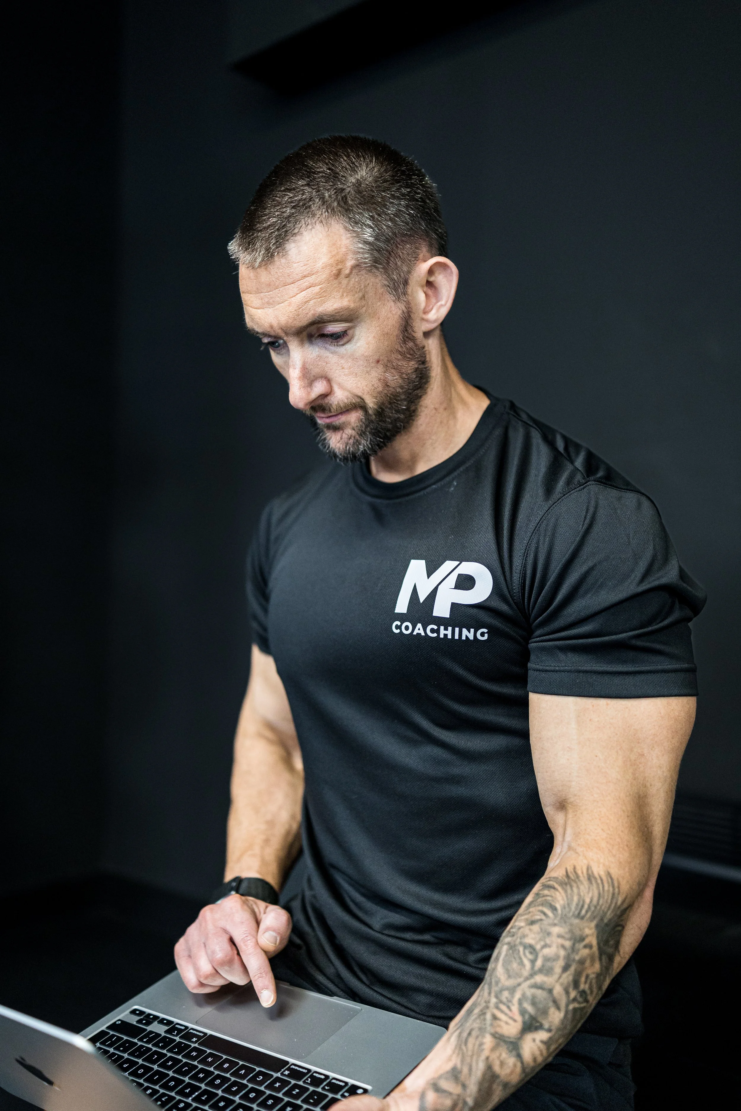 A man with tattoos on his arm wearing a black T-shirt with 'MP Coaching' written on it, looking at a laptop and pointing at the keyboard.
