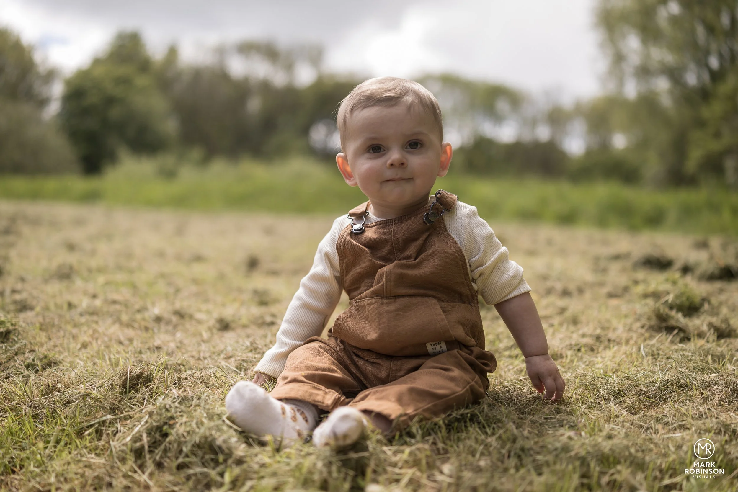 A young boy sitting on the grass outdoors, looking at the camera with a neutral expression, dressed in brown overalls with a cream long-sleeve shirt underneath, in a park or natural setting with trees and cloudy sky in the background.