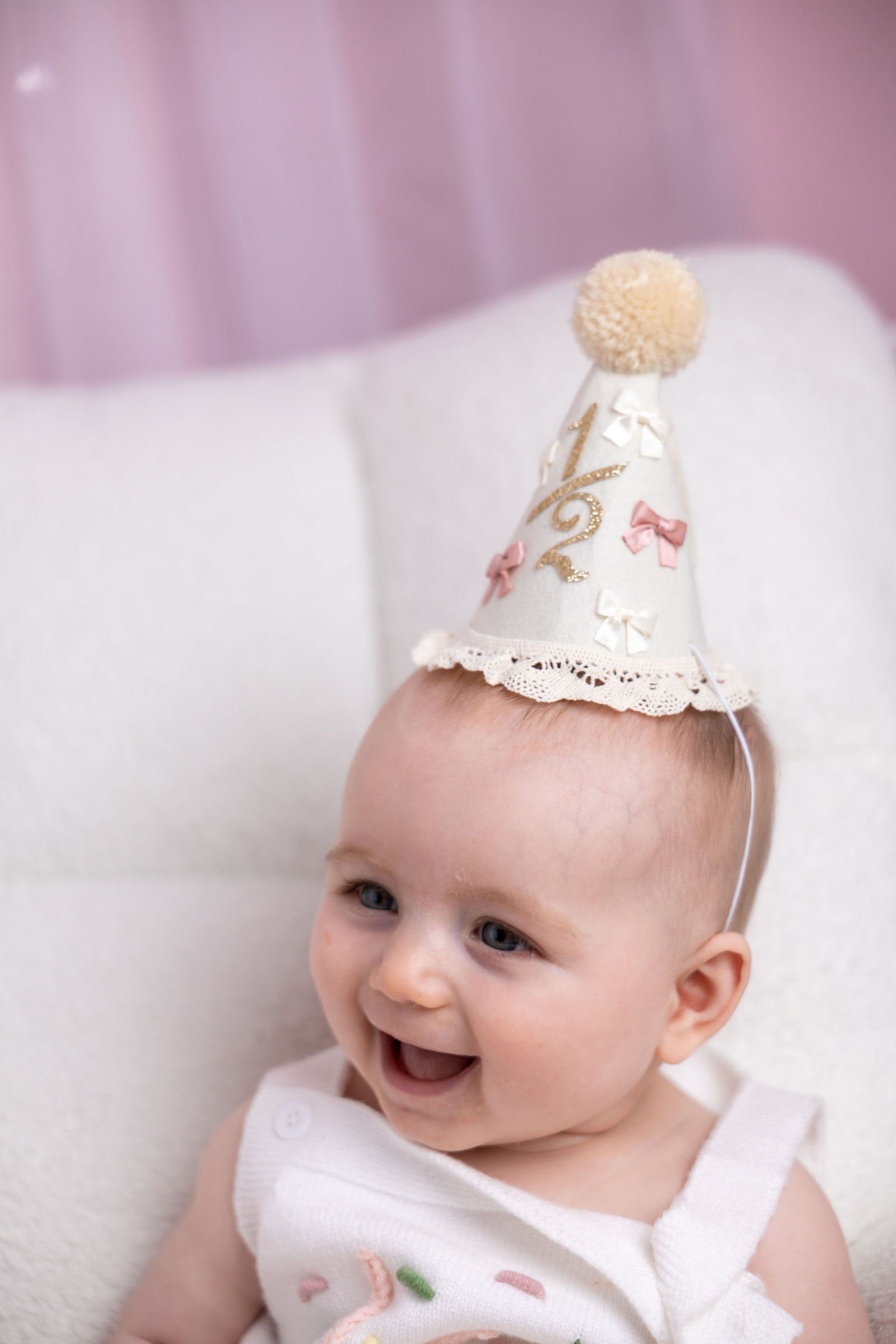 A young child with a joyful expression, wearing a celebratory party hat decorated with bows and ruffles, with a pink pom-pom on top, against a soft pastel background.