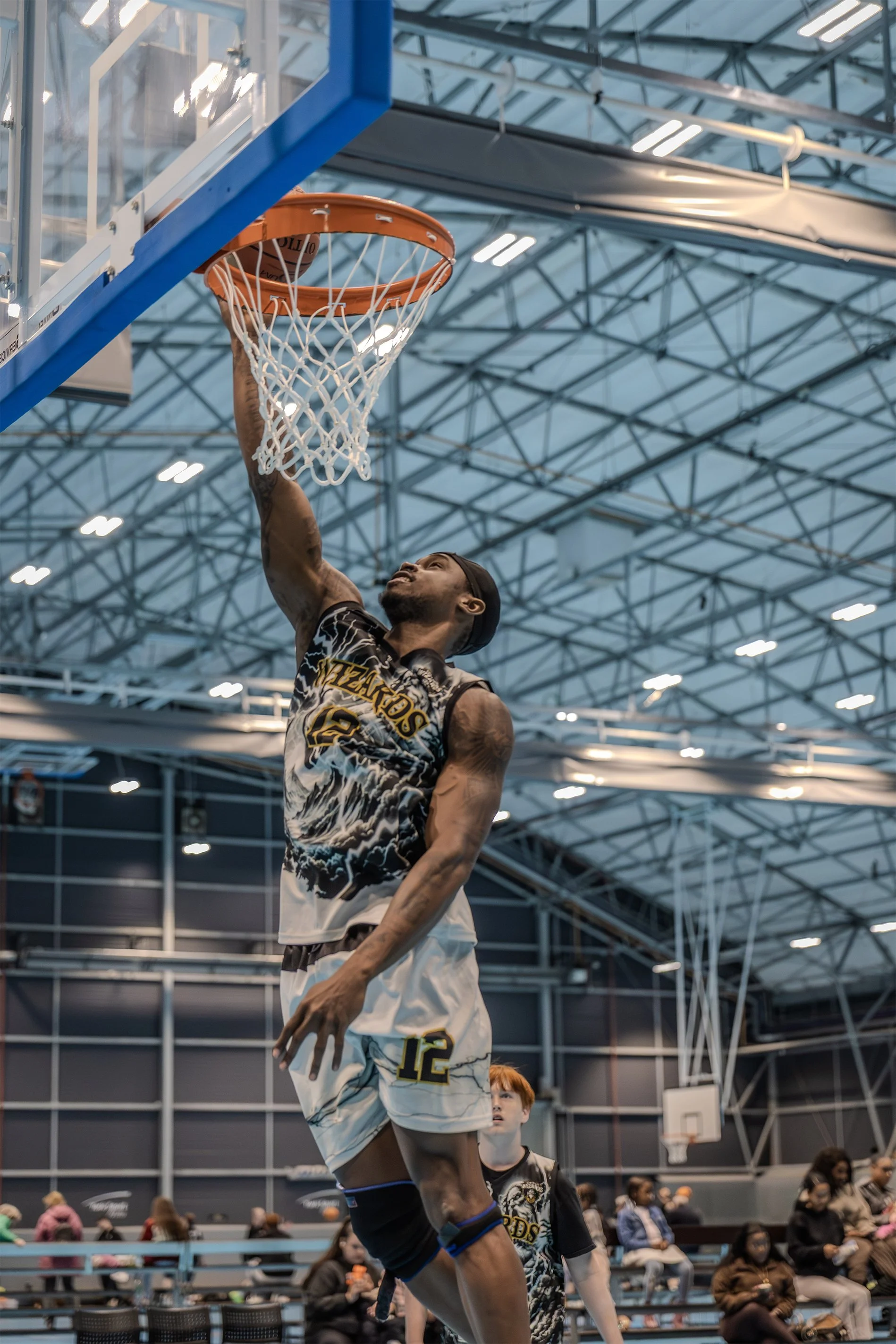 A basketball player in a black and white jersey with the number 12 is jumping towards the basket to dunk the basketball. He is inside an indoor basketball court, with spectators sitting in the background and a high ceiling with lights and structural beams.