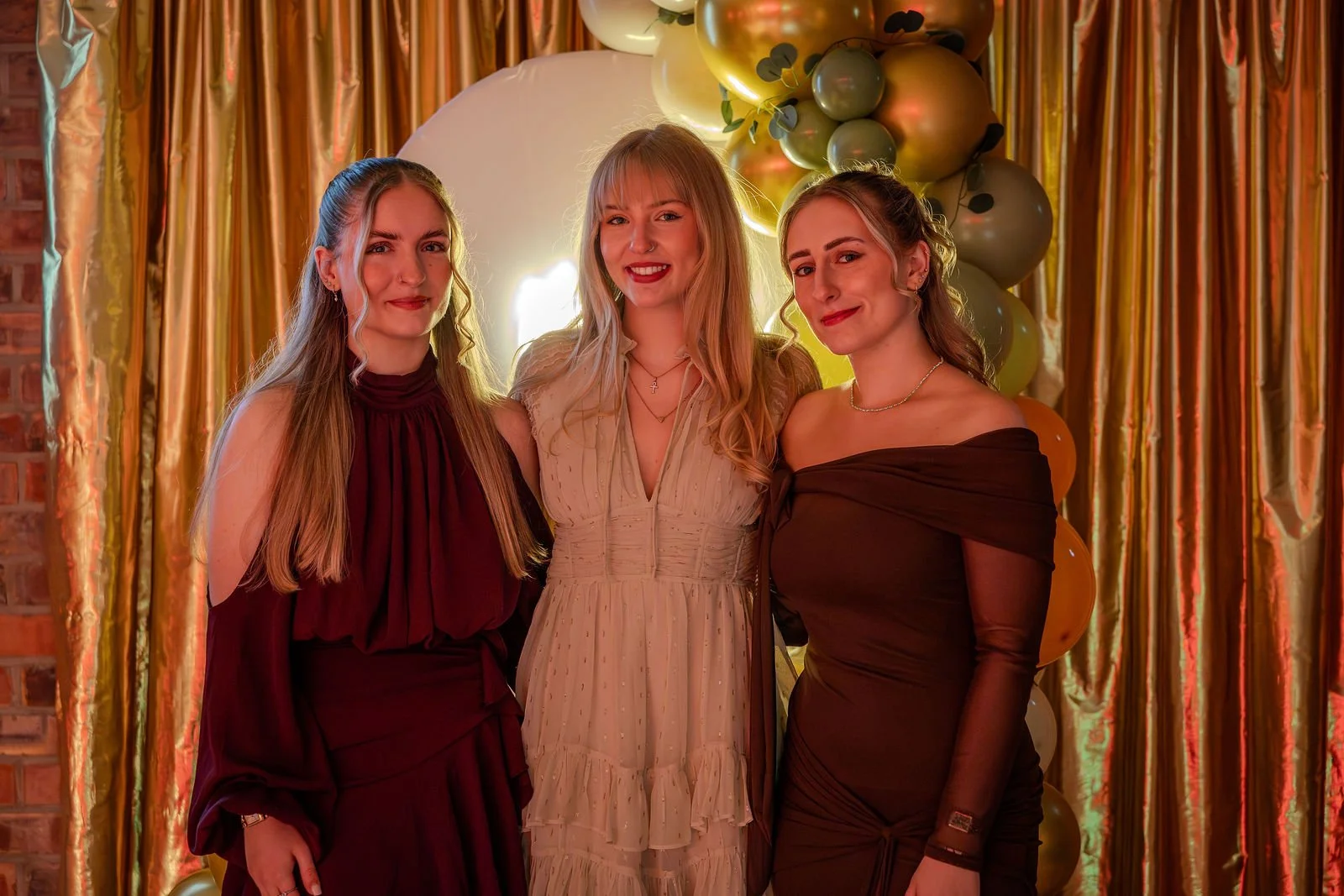 Three women standing together at a celebration event, posing for a photograph in front of a balloon and gold curtain backdrop.