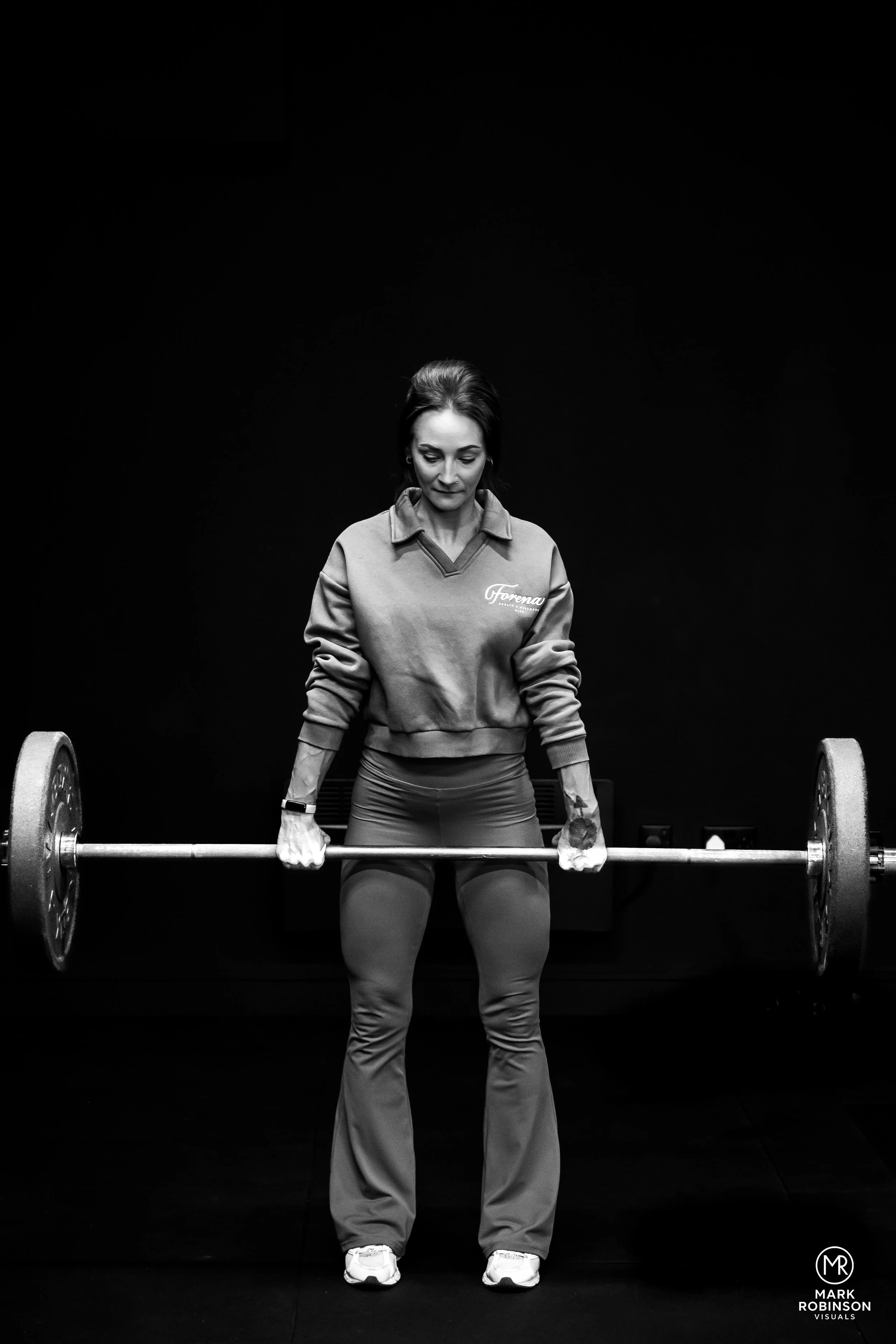 A woman in athletic clothing lifting a barbell with weights in a gym with a dark background.