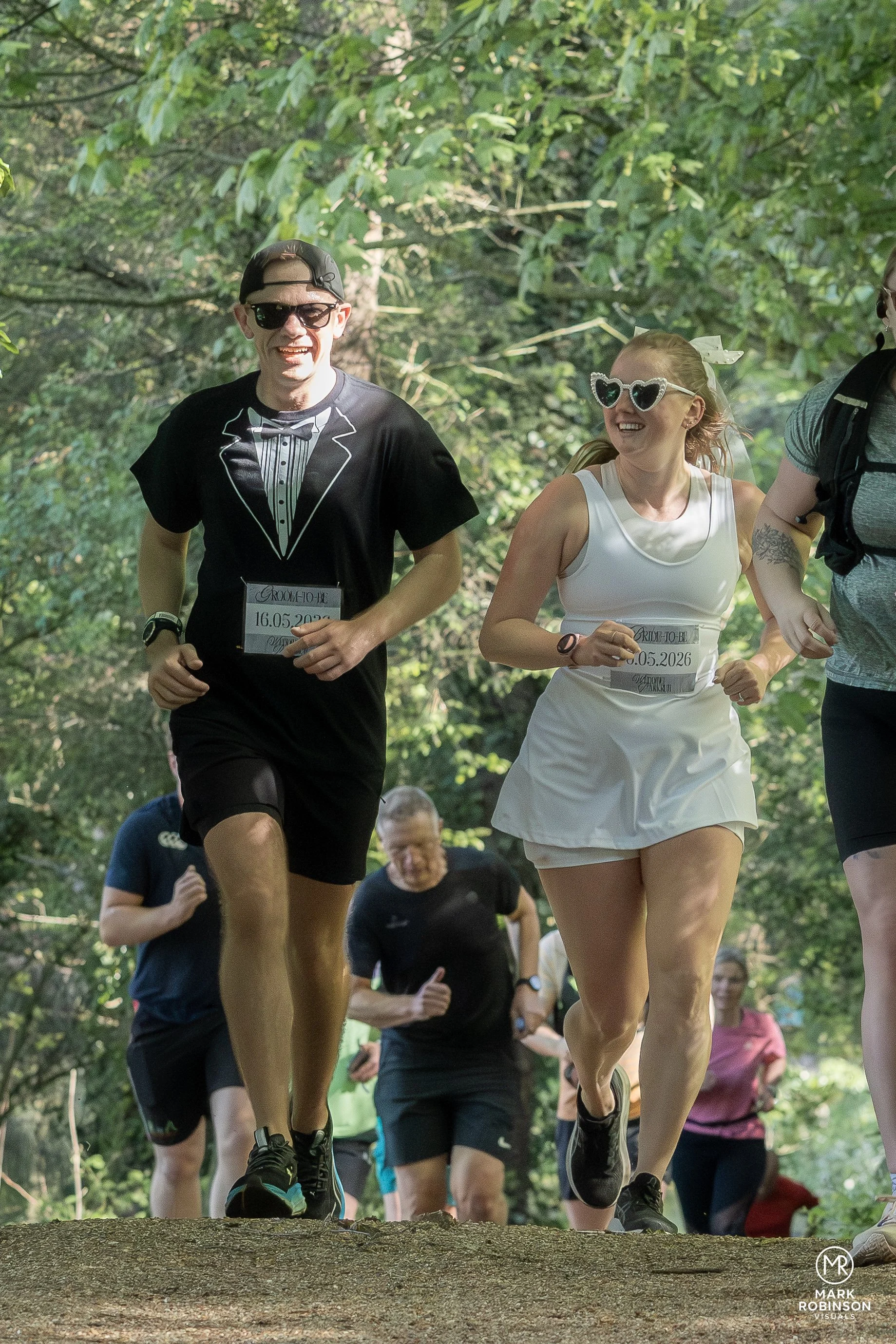 People running in a trail race in a wooded park, wearing athletic clothes and sunglasses, with some bib numbers visible.