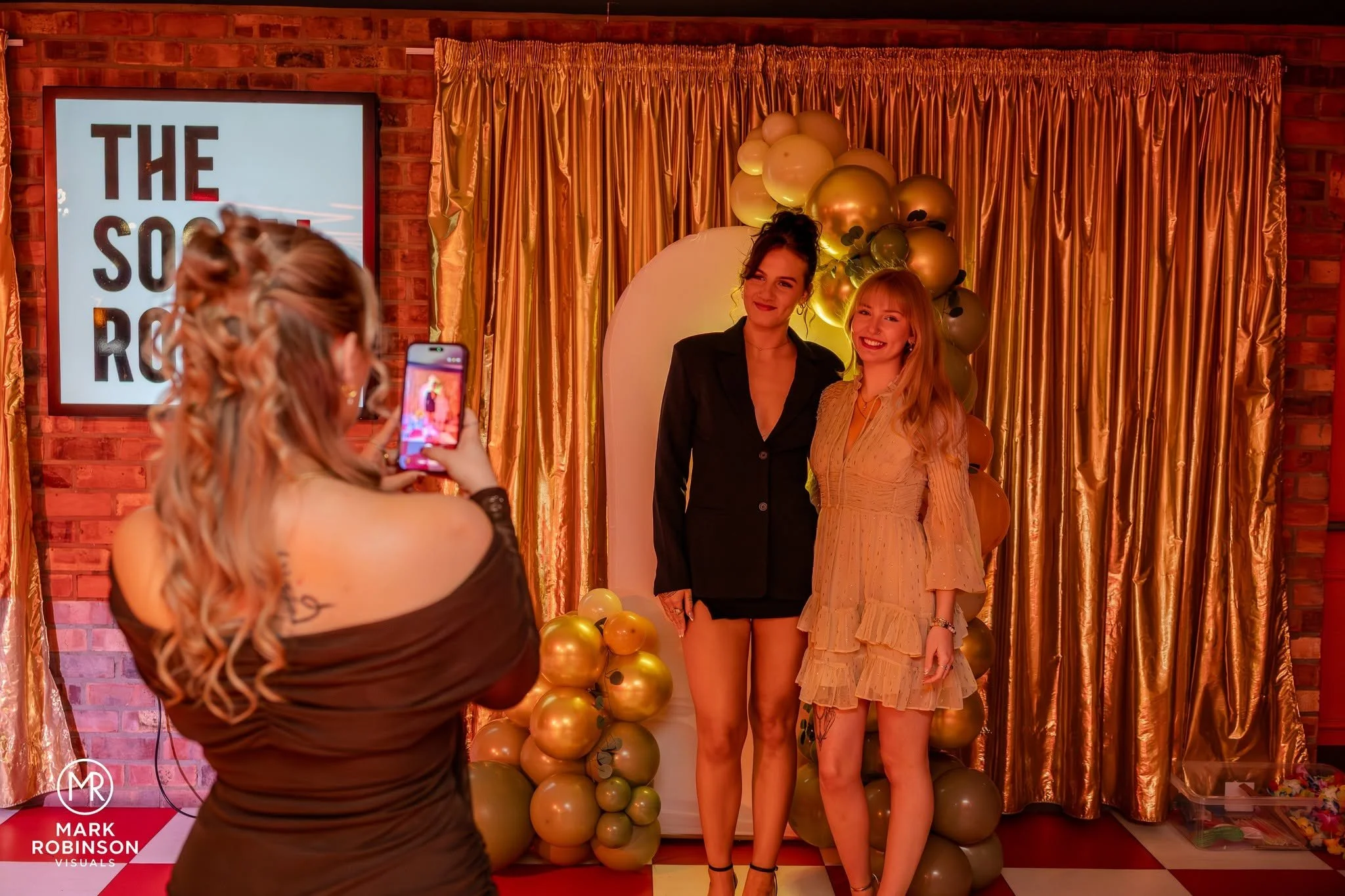 Two women stand in front of a gold curtain backdrop with gold and green balloons at a celebration or event. A third woman is taking their photo with her smartphone. There is a sign on the left side of the image with the text 'THE SOR R' and a logo that says 'MARK ROBINSON VISUALS' at the bottom.