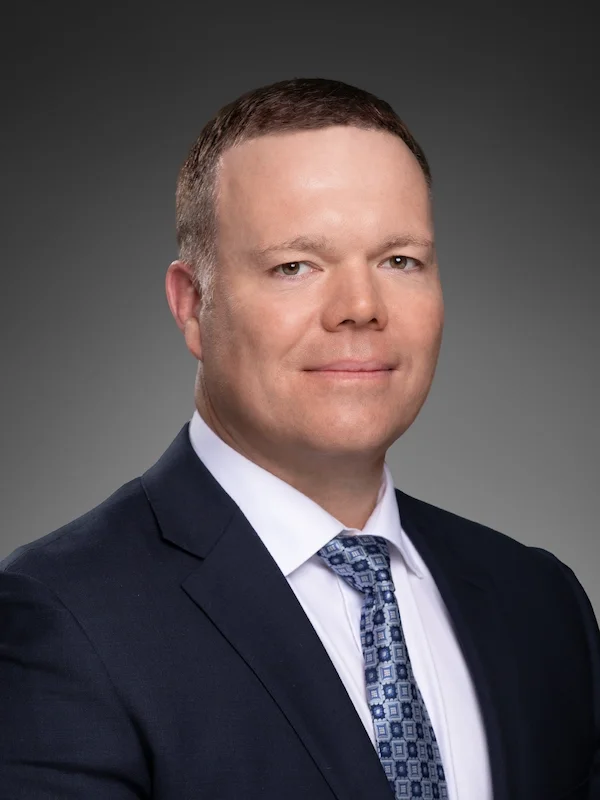 A professional headshot of a man in a dark suit, white shirt, and patterned tie, against a gray background.