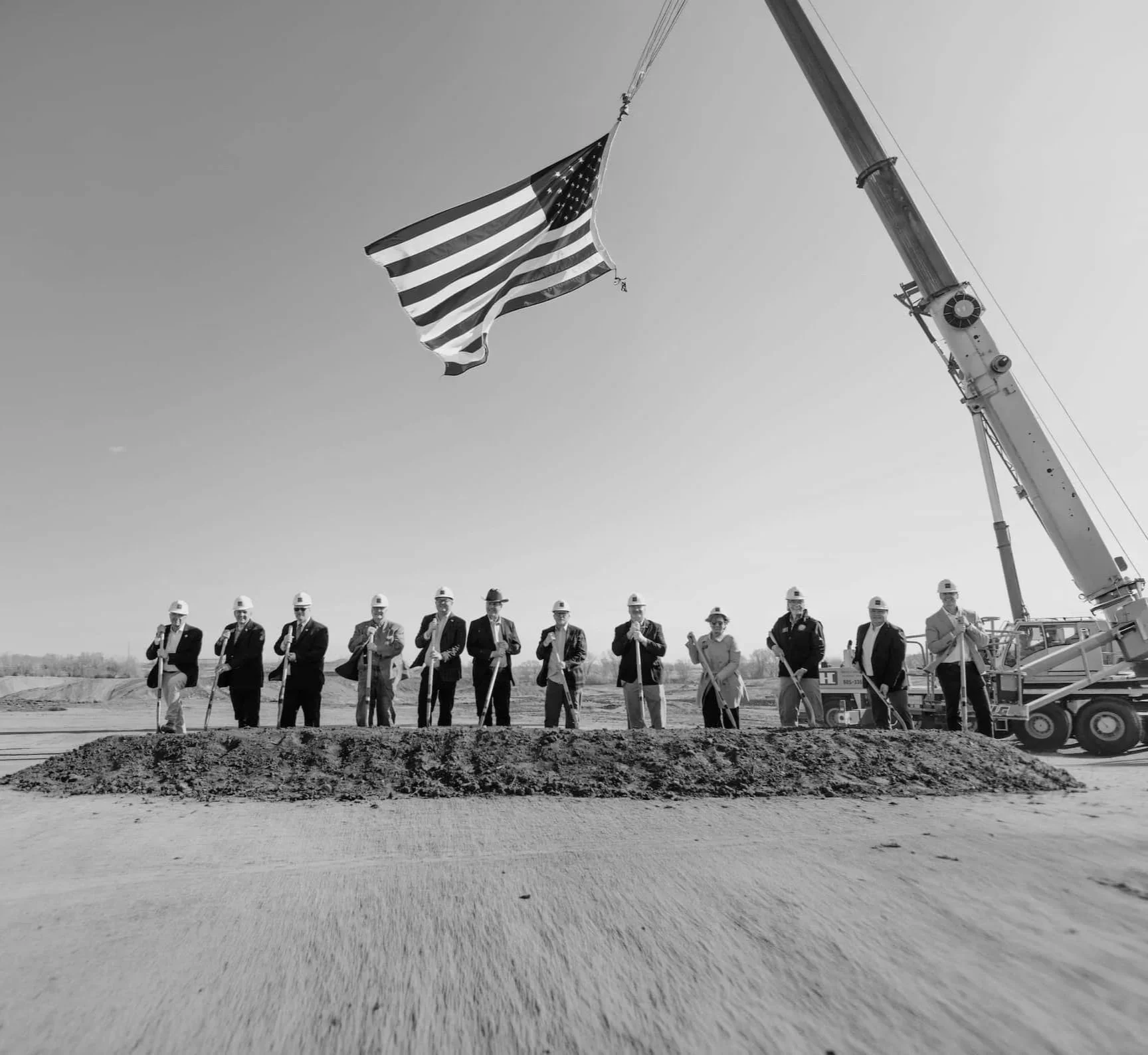 Black and white photo of a group of people in hard hats and work clothes standing on a dirt construction site holding shovels, with a large crane lifting an American flag overhead.