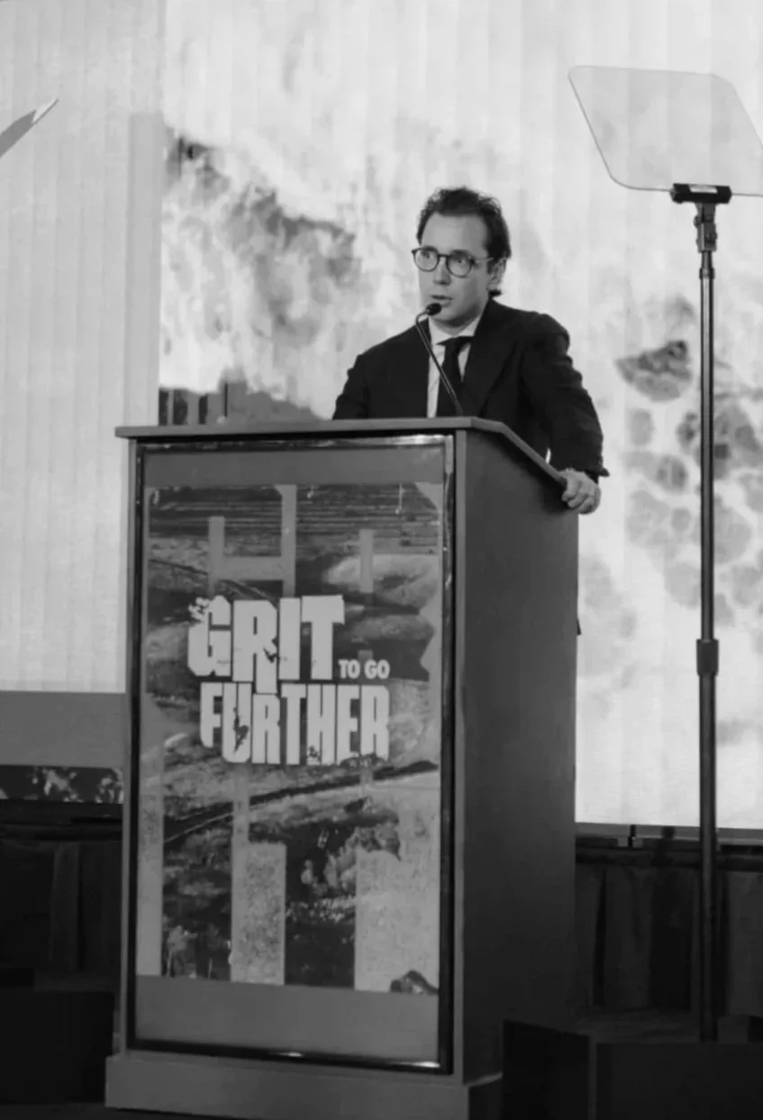 A man with glasses and a suit standing at a podium giving a speech, with a large screen behind him displaying an abstract background. The podium features the logo for the video game 'Grit to Go Further'.