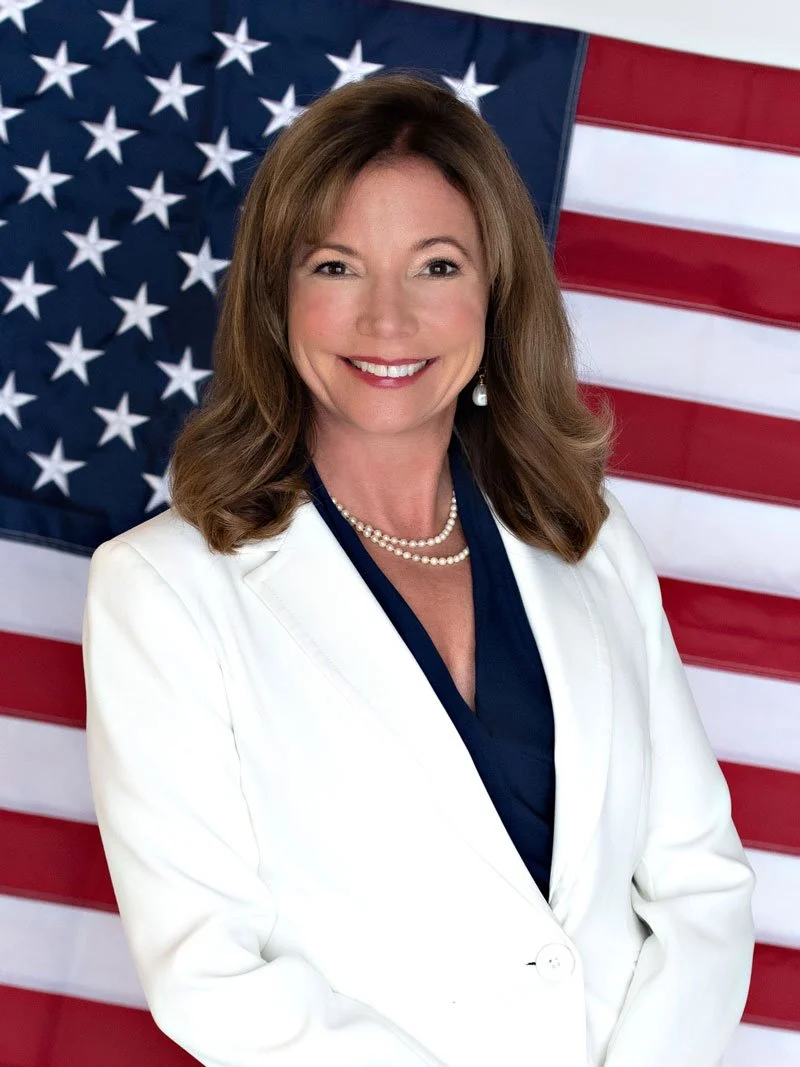 A woman with shoulder-length brown hair smiling in front of an American flag, wearing a white blazer, navy blouse, pearl necklace, and pearl earrings.