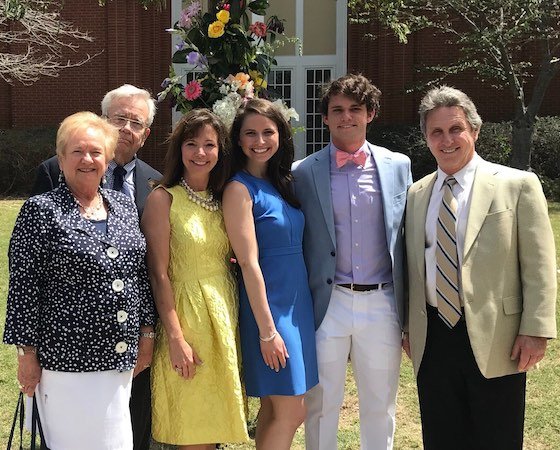 Group of six people outdoors, three women and three men, posing for photo in front of flowers and a building, dressed in summer clothing.