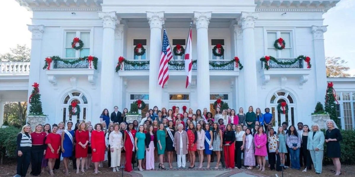 Group of women and a few men dressed in formal attire standing in front of a white building decorated with red and green Christmas wreaths and bows, with American and state flags hanging from the columns.