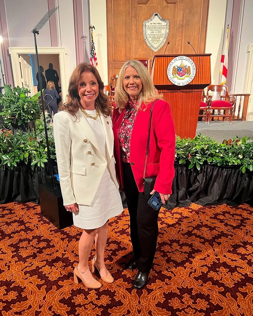 Two women standing together indoors in front of a podium with a seal that says 'County of the Gore of the City of Allen.' One woman is wearing a white suit with a skirt and heels, and the other is wearing a red blazer with black pants and boots. There are chairs, plants, and flags behind them.