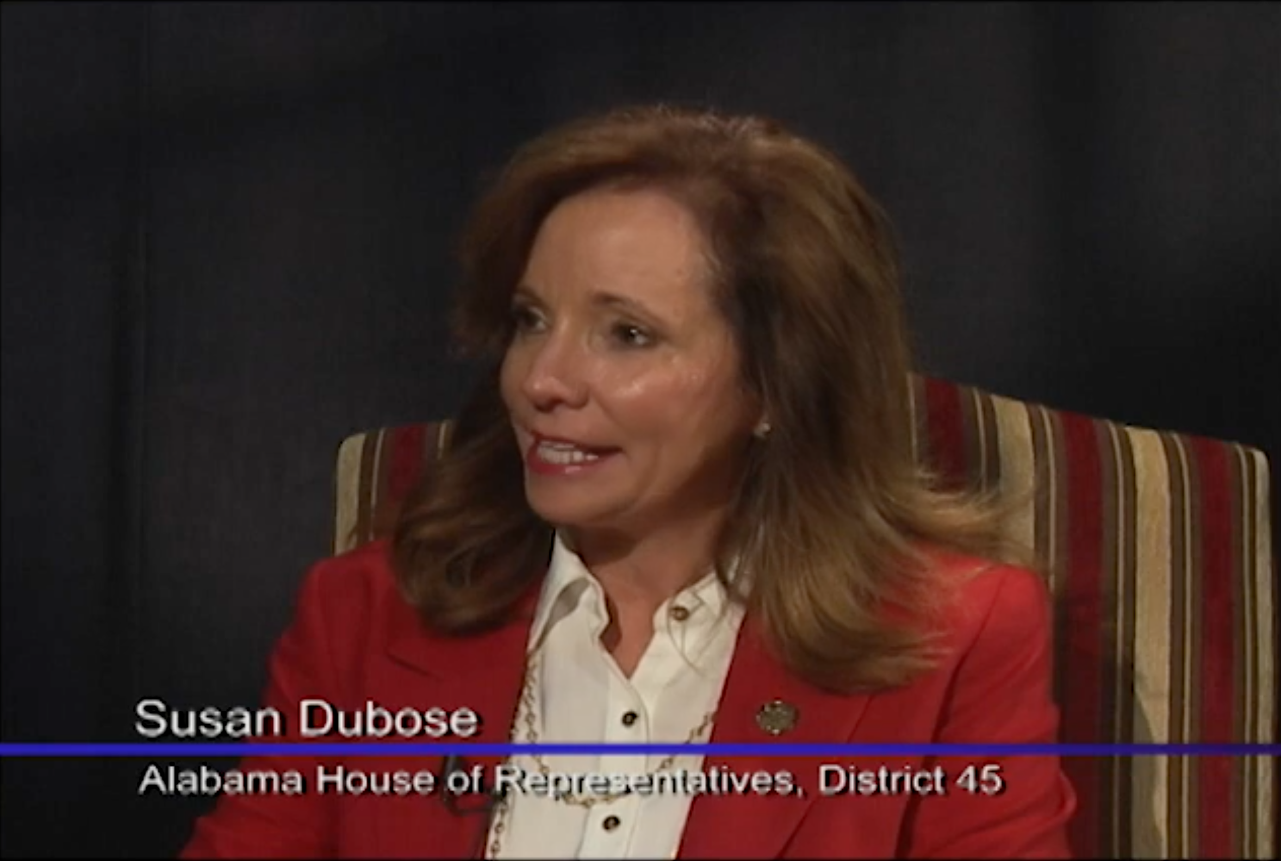 A woman with brown hair wearing a red blazer and white blouse, sitting on a striped chair against a dark background. Text overlay identifies her as Susan Dubose from Alabama House of Representatives, District 45.