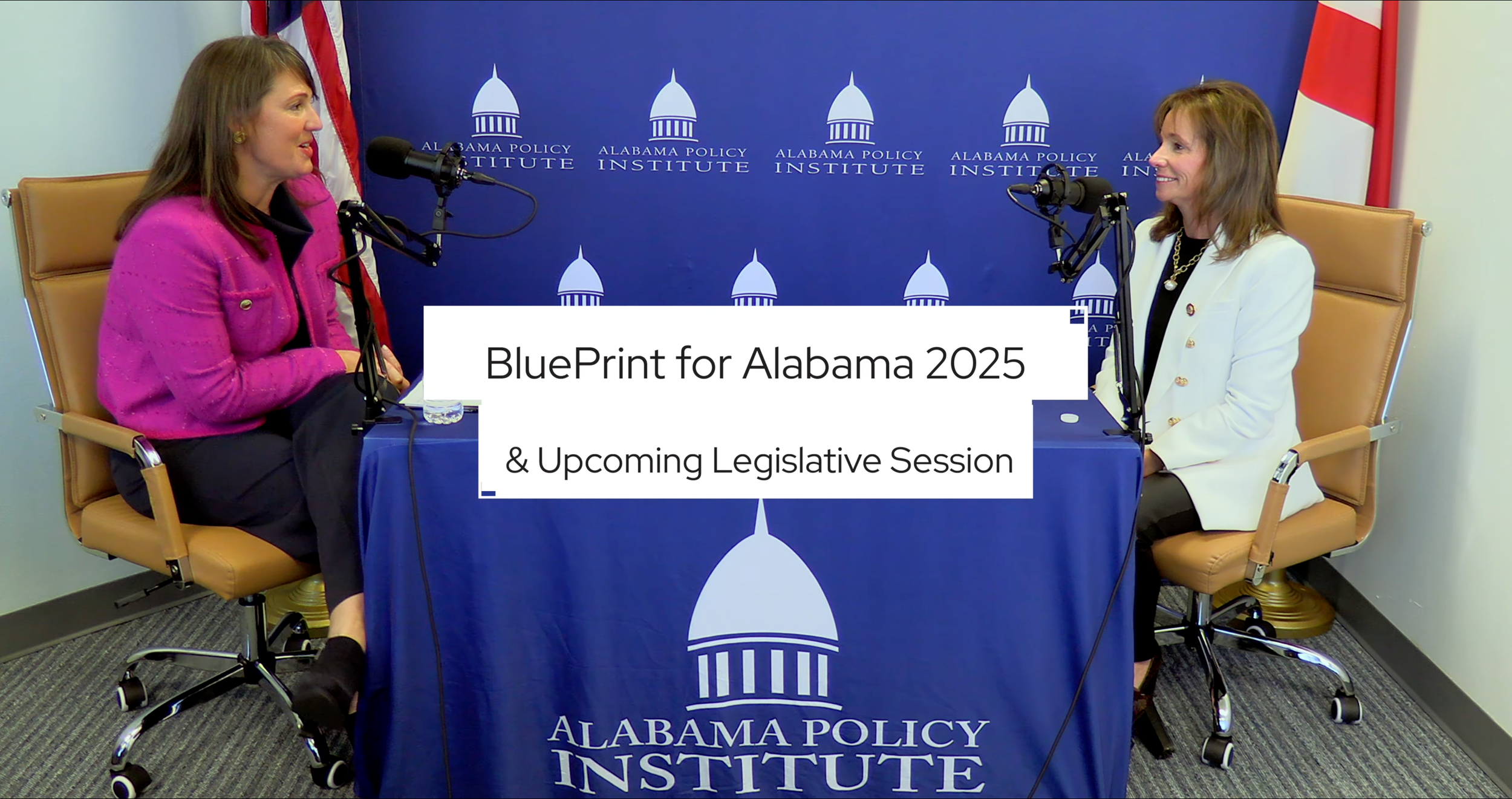 Two women seated at a table with microphones, engaged in conversation, in front of a backdrop with the Alabama Policy Institute logo. The table has a blue cloth with the organization's logo and text.