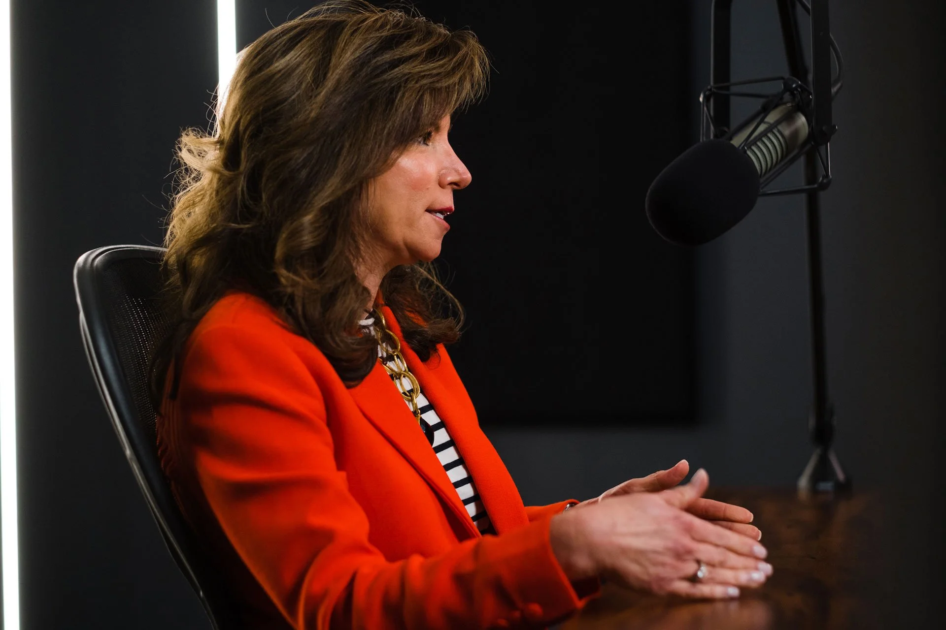 A woman with brown hair wearing an orange blazer and striped shirt, sitting at a desk in front of a microphone in a recording studio.