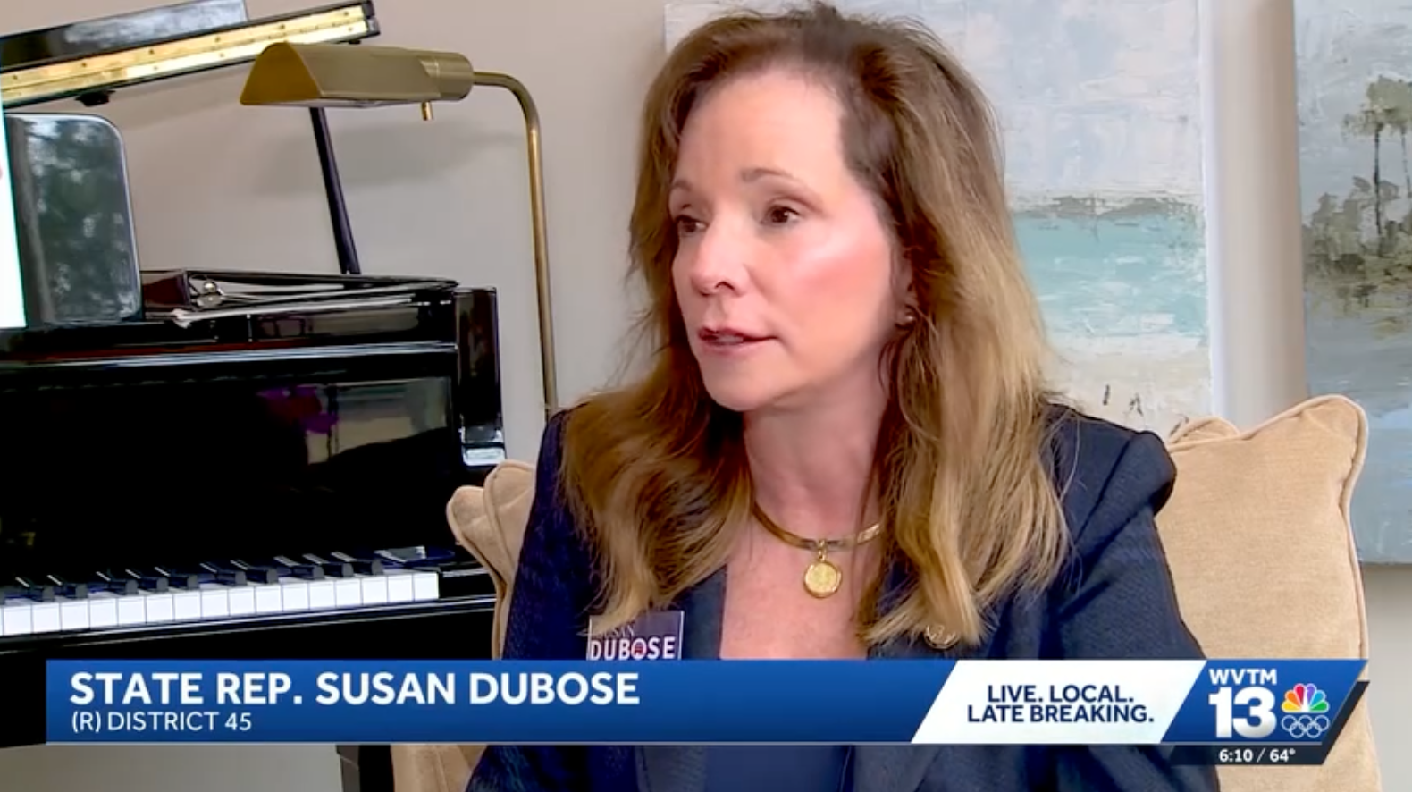 A woman with shoulder-length reddish-brown hair wearing a dark blazer and a gold necklace, sitting in a beige chair, being interviewed during a TV news broadcast. Behind her is a black upright piano with a music stand and a pixelated painting with light blue, white, and gray tones. A blue banner at the bottom of the screen displays her name as State Rep. Susan Dubose, and her district information, along with news station details.