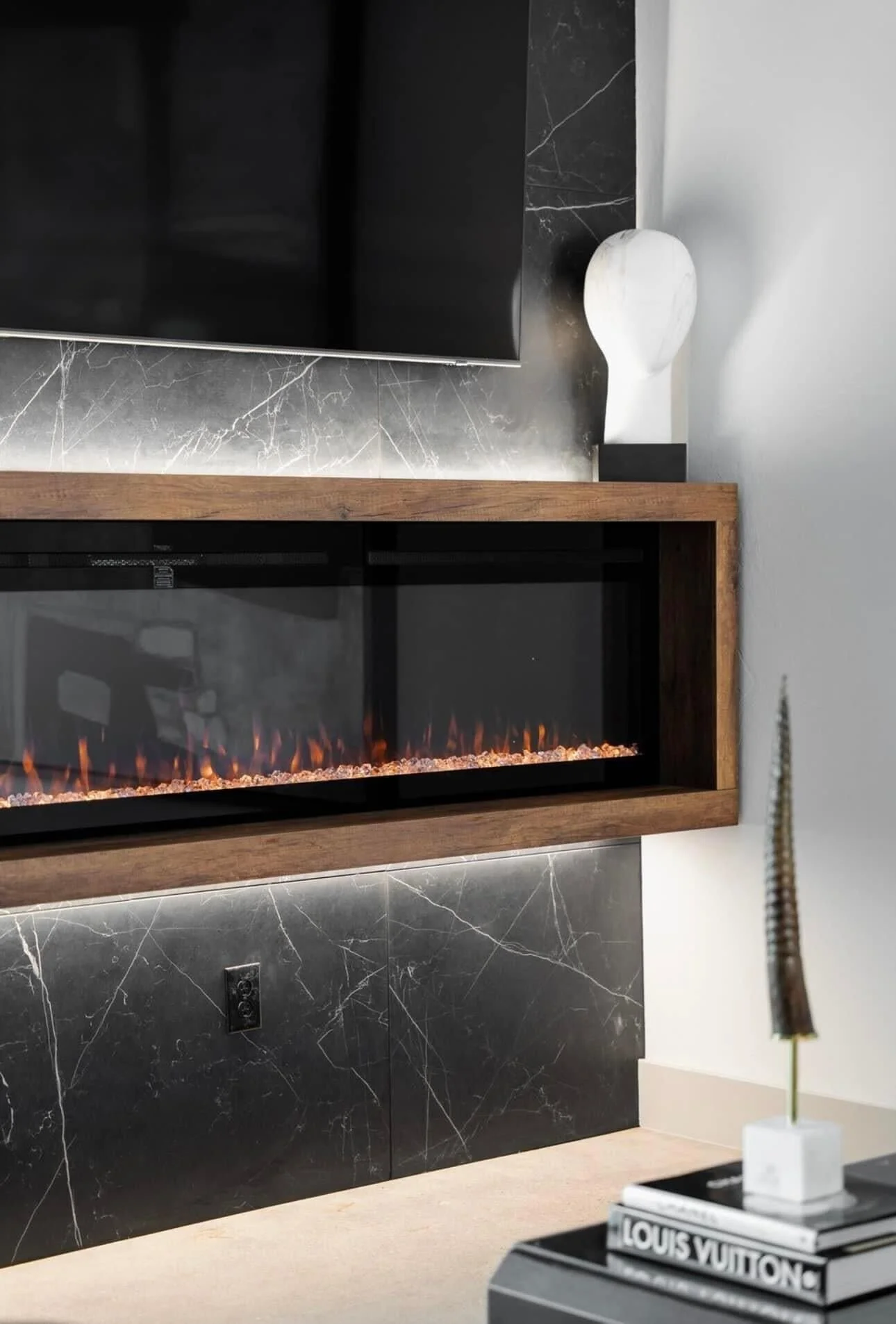 Modern living room with a black marble wall, a wall-mounted electric fireplace with a wooden frame, and a white decorative object on a shelf. Some books and a flower are also visible.