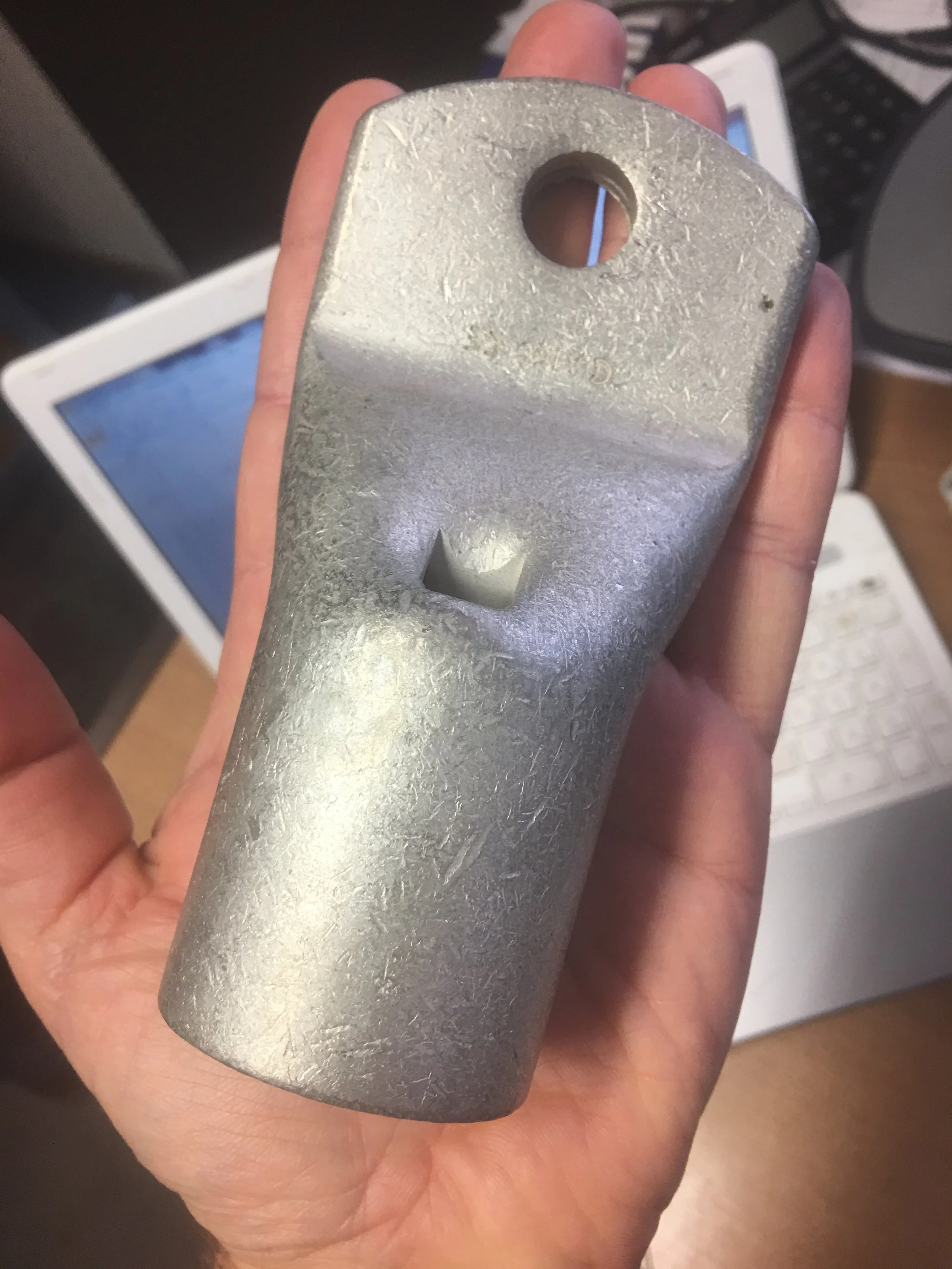 A hand holding a metallic device with a hole at the top and a small square opening in the middle, set against a background with a laptop and a desk organizer.