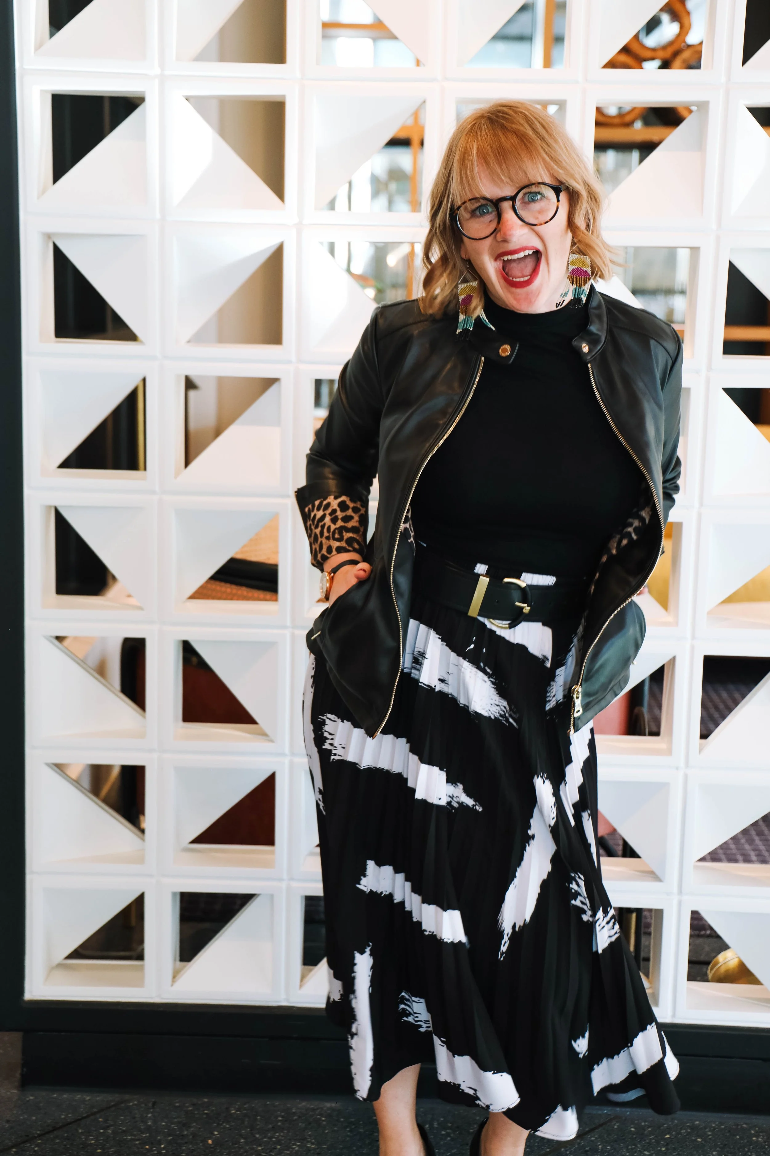 kari swinging hips in front of geometric white patterned wall cutout wearing black and white skirt leather leopard print lined jacket, colorful earrings, red hair, glasses, bangs, red lips with a big open smile