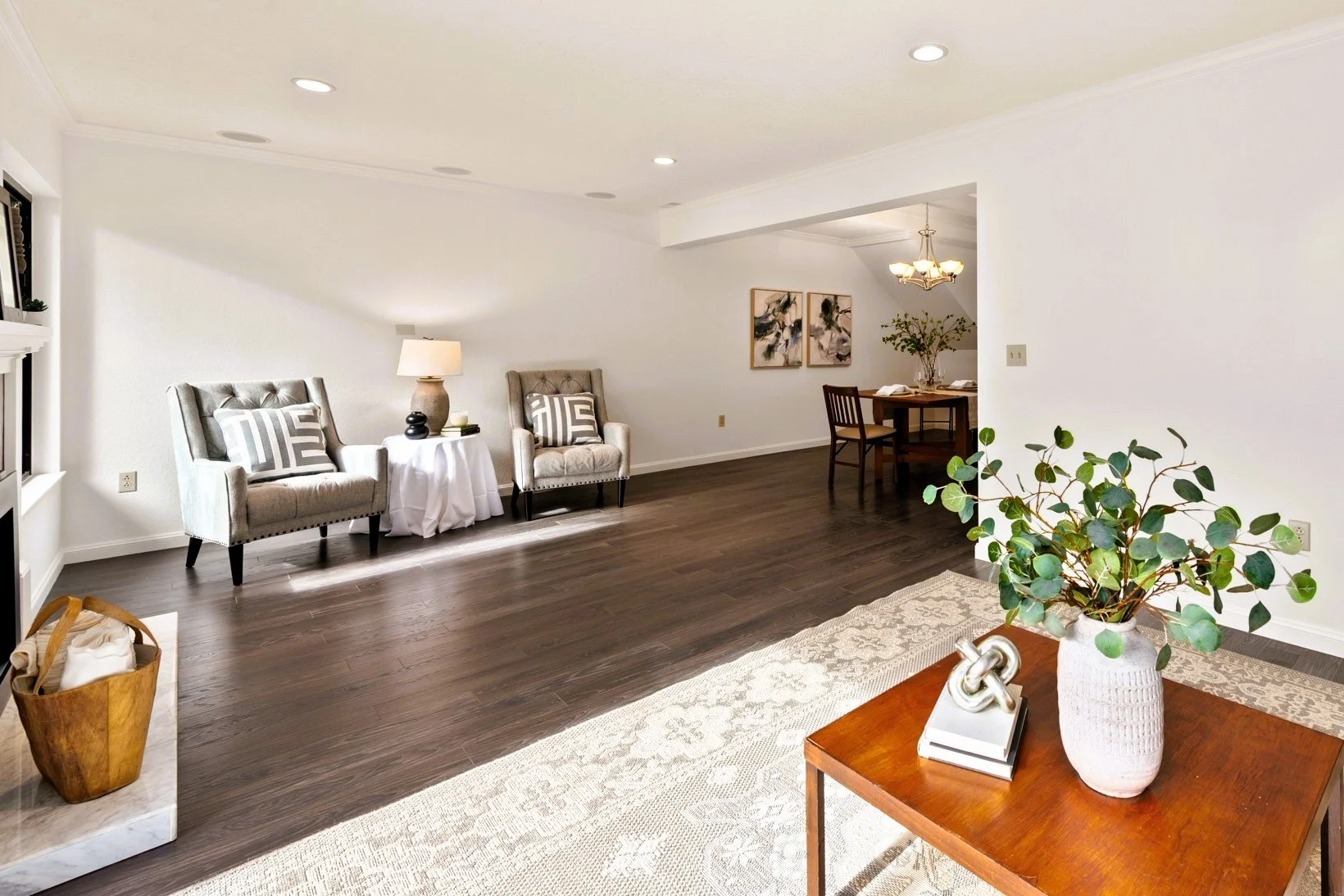 Bright living room with dark hardwood floors, two beige armchairs with patterned pillows, a small round side table with a lamp, and a large area rug. A wooden table with greenery and decorative items is in the foreground, with a sitting area and dini