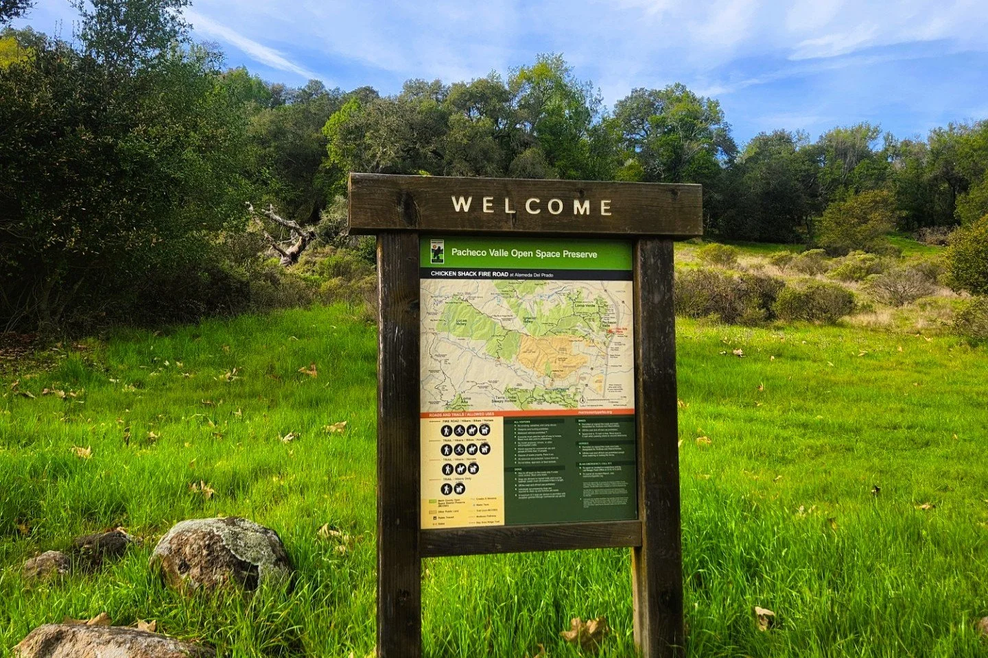 A wooden welcome sign with a map at the Pacheco Valle Open Space Preserve, surrounded by green grass and trees under a blue sky.