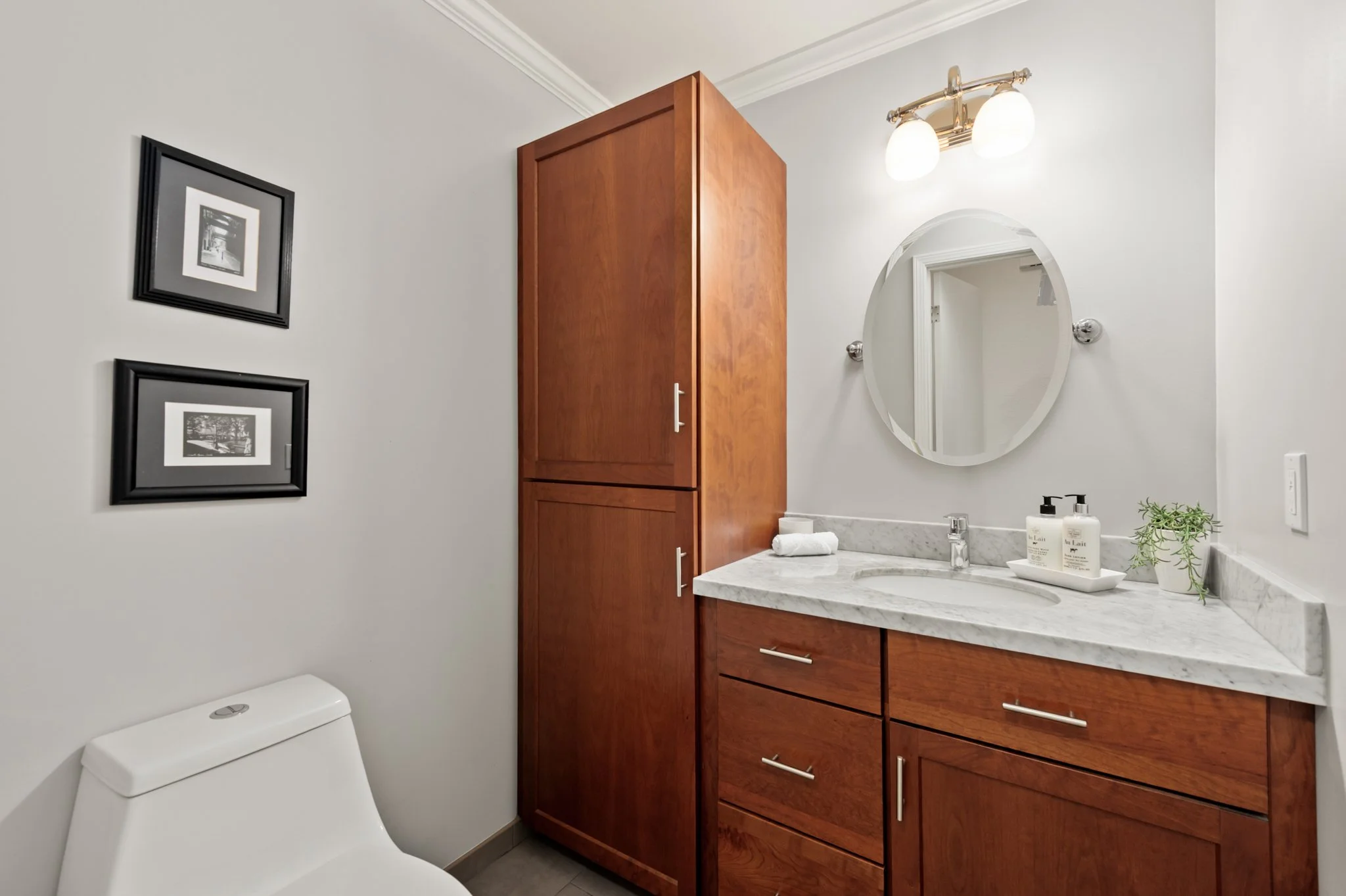 A modern bathroom with a white toilet in the lower left corner, a wooden cabinet and vanity with a marble countertop, a round mirror, and a light fixture with two globes above the mirror. There are two framed black-and-white photographs on the wall.