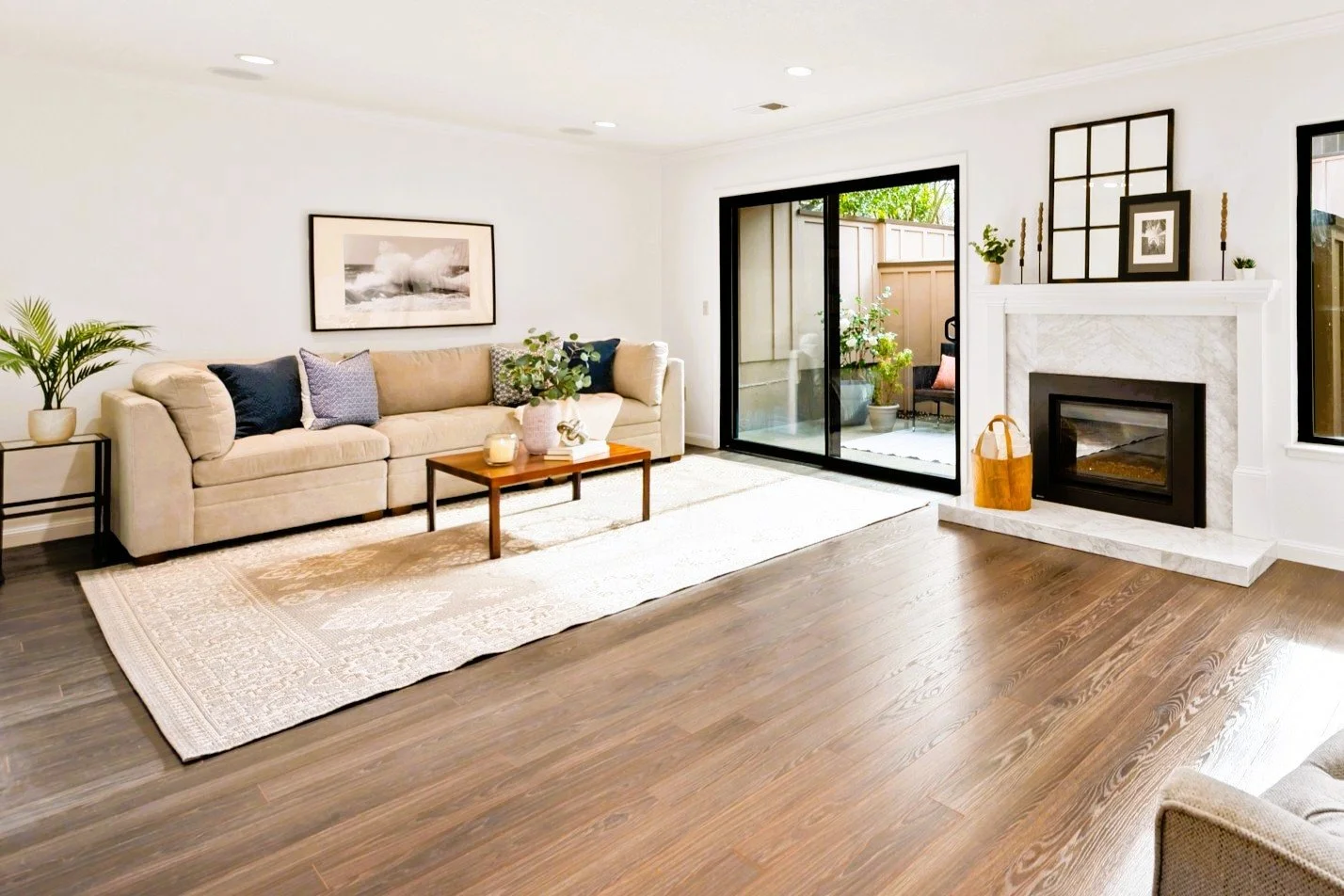 Living room with cream sofa, wooden coffee table, fireplace, hardwood floor, sliding glass door leading to patio, potted plants, and wall art.