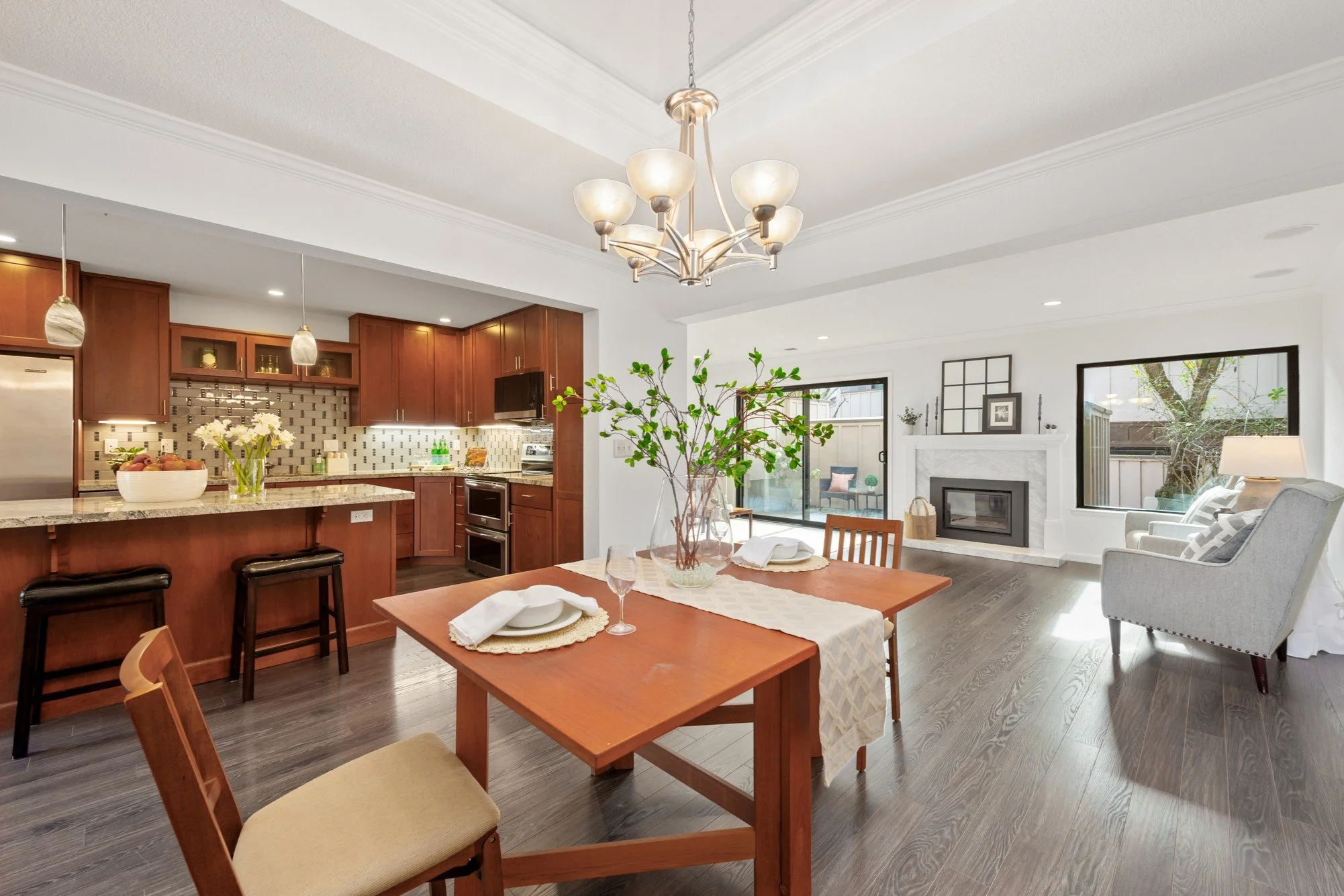 Open-concept living and dining room with kitchen, wooden dining table set for two, a gray armchair, white fireplace, sliding glass door, and large windows allowing natural light.